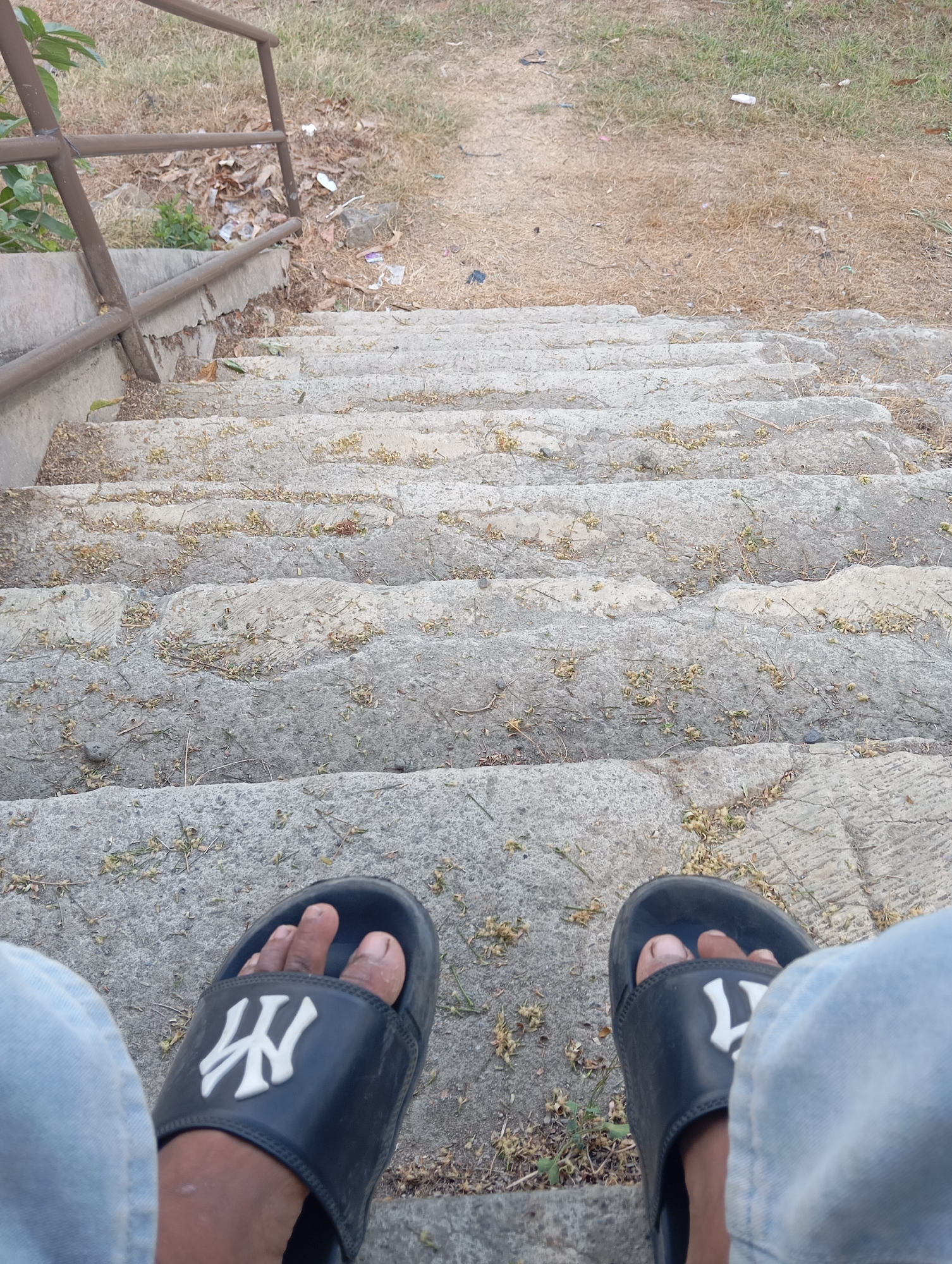 The image shows a first-person perspective of a person sitting on stone stairs outdoors, likely in Sokaraja, Indonesia. The person is wearing blue jeans and black slides with a white "NY" logo. The stairs are worn and covered in small debris, possibly dried leaves or flower petals. To the left of the stairs, a metal handrail is visible, and beyond that, dry grass and some scattered litter. The background consists of dry vegetation and a path leading upwards.