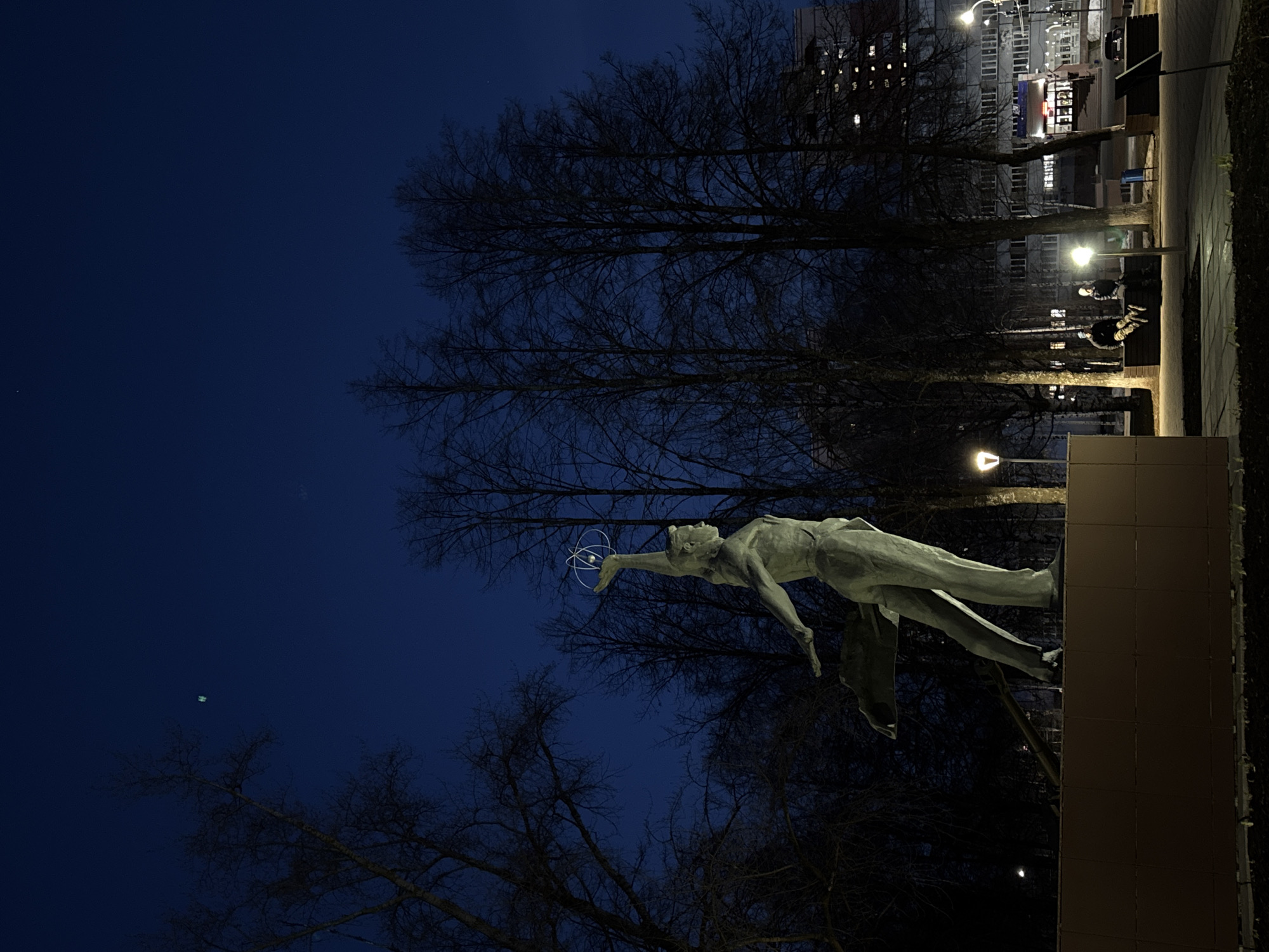 The image shows a statue, "Friendship of Peoples", in Izhevsk, Russia, at night. The statue depicts a man holding a model of an atom. It is mounted on a rectangular base of brown tiles. In the background, there are bare trees and illuminated buildings. There are also lampposts providing light to a pathway. A couple is visible sitting on a bench in the background. The sky is a deep blue.