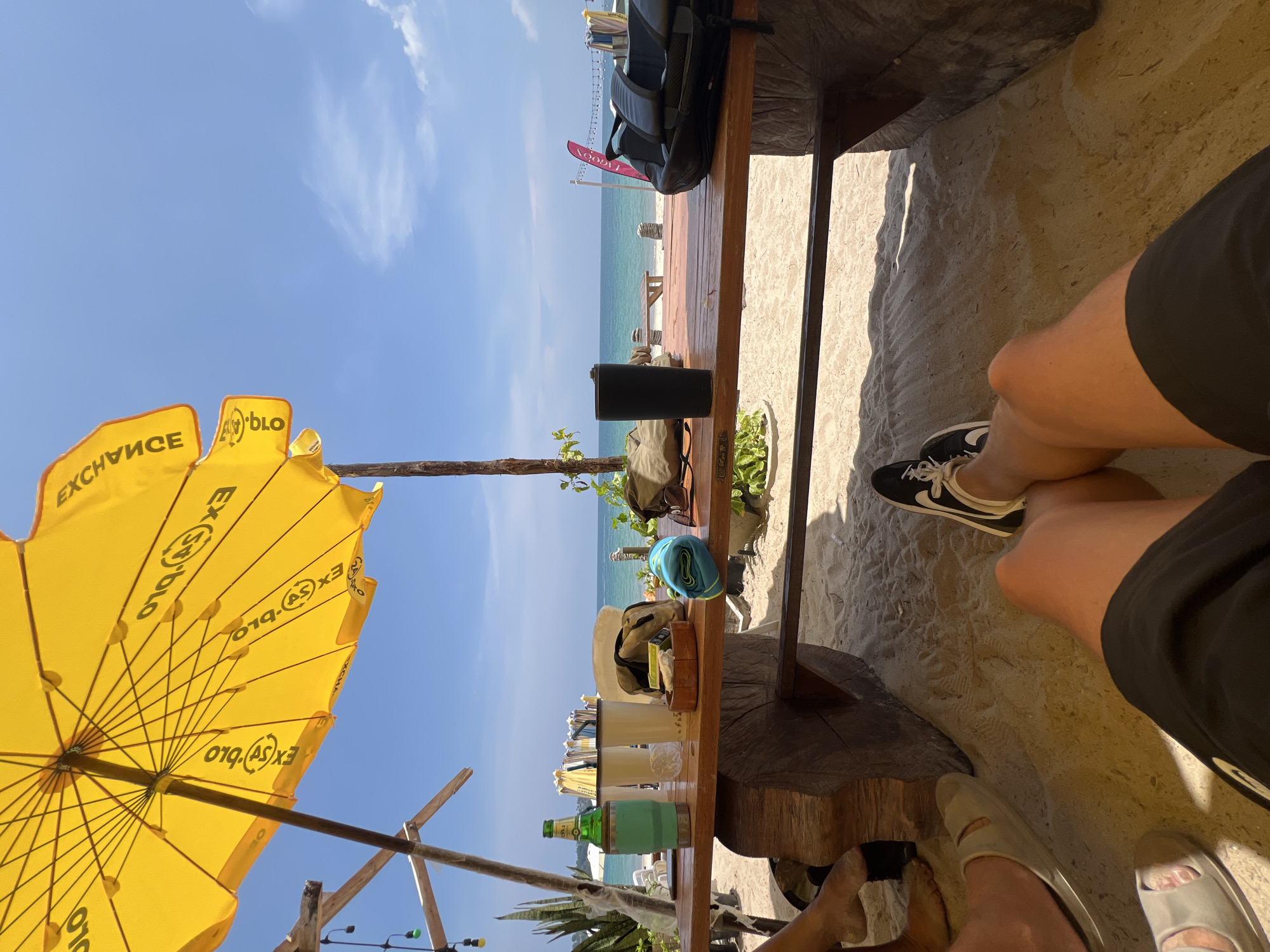 The image captures a vibrant beachside scene from a first-person perspective, looking out from under a yellow umbrella towards the ocean. The viewer appears to be seated at a wooden bar or table, with sand underfoot.
In the immediate foreground, the legs and feet of at least two individuals are visible. One person, on the right, wears black shorts and black sneakers with white laces, featuring a white Nike swoosh logo. Another person's feet, wearing light-colored slip-on sandals, are partially visible on the left.
The wooden table, or bar surface, stretches across the middle of the frame, laden with various items. These include a black portable speaker, a tall green bottle (possibly beer or a spirit), several white plastic cups, a teal insulated tumbler, sunglasses, a hat, and a small wooden bowl containing a salad or green food item. Beyond the table, the bright blue-green ocean stretches to the horizon under a clear, sunny sky with sparse white clouds. In the distance, other beach umbrellas and wooden structures are visible on the sand near the water. A red banner or flag, partially obscured, stands on the beach, displaying the reversed text "LAGOON".
Dominating the left side of the frame is a large yellow beach umbrella, providing shade. The umbrella's fabric features visible, reversed text: "EXCHANGE" and what appears to be "EX(S)BIO". Wooden support beams and string lights are also visible around the seating area. The bright sunlight suggests it is daytime, likely mid-morning or afternoon, and the overall atmosphere is one of relaxation and leisure by the sea.