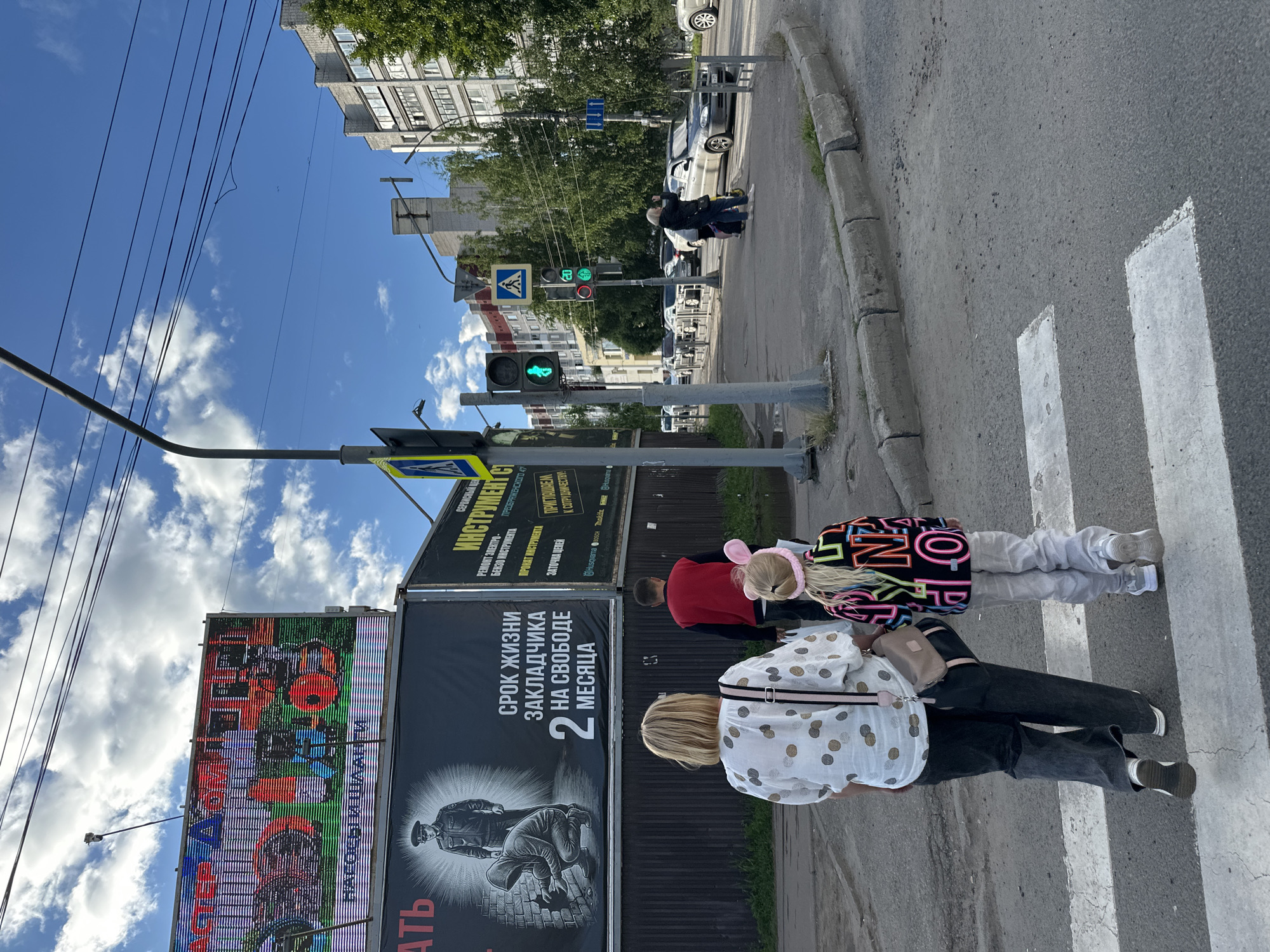 The image shows a street scene in Rubtsovo, Russia, on a sunny day with scattered clouds. The perspective is from behind a group of people walking on a crosswalk, looking towards the street and buildings.
In the foreground, three people are visible from the waist down, walking on a zebra crossing. The person closest to the camera is wearing a white shirt with a polka dot pattern and dark pants. The middle person is wearing dark pants and a light-colored shirt. The person furthest away is wearing a black jacket and possibly dark pants. Two of them have bags.
The scene depicts a street with a sidewalk, a paved road, and buildings in the background. Traffic lights are visible, with one showing a green pedestrian signal. A large billboard with colorful advertising is on the left side of the image. Another billboard displays text in Russian. The general atmosphere appears to be one of everyday urban life. The time of day is likely daytime, given the bright sunlight and clear sky. There are no obvious indications of any specific event or strong emotions. The text on the billboards is in Russian, with one legible phrase appearing to be "Срок жизни закладчика на свободе 2 месяца," which translates to "The jail time for a drug dealer is 2 months."