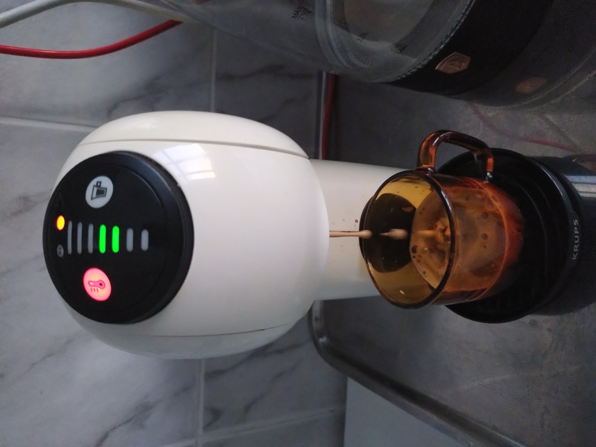 The image shows a Krups Dolce Gusto coffee machine dispensing coffee into a brown glass mug. The machine is white with a black control panel featuring indicator lights, and it is attached to a water tank. The background includes a white tiled wall and a red cable. The location appears to be indoors, likely in a kitchen or pantry. It is not possible to determine if the image was taken in Stara Zagora, Bulgaria from this image alone.