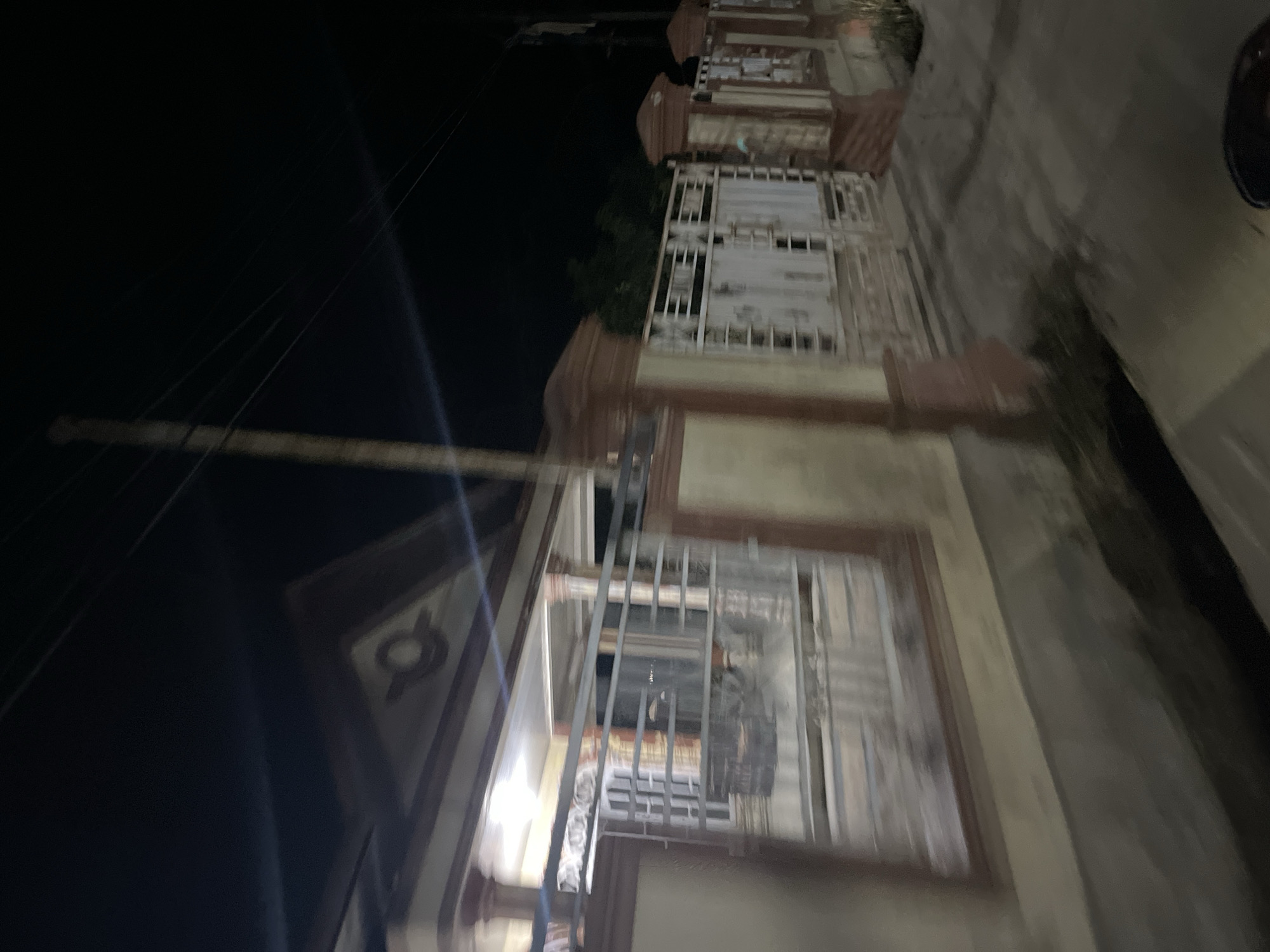 The image depicts a dimly lit scene at night, likely in Menanggini, Indonesia. The primary subject is a building with an ornate facade, featuring multiple levels and windows with metal grates. The building appears to be constructed with brick or concrete, accented with reddish-brown trim. The windows are mostly covered or obscured, suggesting an unoccupied or perhaps secured space.
The perspective is from street level, looking upwards and across the building. The overall impression is one of quiet and perhaps a bit of mystery due to the low light conditions and the barred windows. There is a sense of depth created by the receding lines of the building and the street.
The scene is set at night, as indicated by the darkness and the artificial lighting that casts faint glows and reflections. The weather appears to be clear, with no visible rain or wind effects. There are no people or animals visible in the frame, adding to the stillness of the environment.
Notable details include the architectural style of the building, which suggests a historical or traditional design. The presence of what appear to be utility lines or wires running diagonally across the upper left portion of the image adds a touch of everyday realism to the scene. The blurriness of some parts of the image, particularly in the foreground, suggests movement of the camera or a low light setting causing a longer exposure.
There is no visible text in the image. The overall mood is contemplative, with the darkness and the imposing architecture creating a somber or intriguing atmosphere. The location context of Menanggini, Indonesia, places the scene within a specific cultural and geographical setting, though without further information or clearer visual cues, specific cultural markers are difficult to identify.