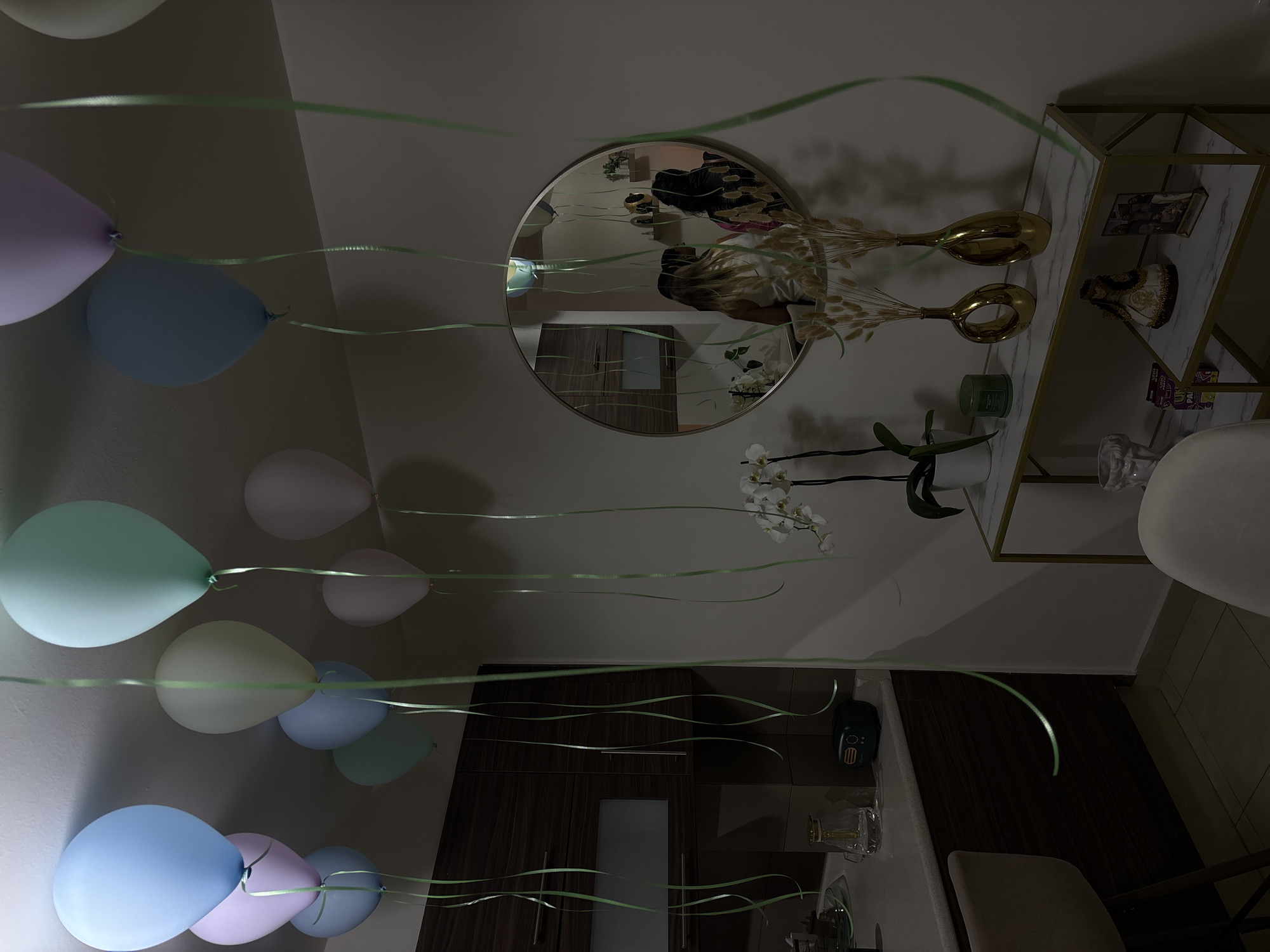 The image depicts an interior space in what appears to be a residence. The walls are light in color, and the lighting is dim. Several balloons in pastel shades of blue, purple, pink, and white are suspended from the ceiling by green ribbons. A round mirror reflects a person taking the photo. A shelf with gold accents displays decorative items, including two golden vases, a figurine, and other miscellaneous objects. A white orchid is also visible. The lower portion of the image shows dark wooden cabinets, part of a countertop, a vacuum and the back of a chair. The location cannot be confirmed to be in Puebla, Mexico based on the image alone.
