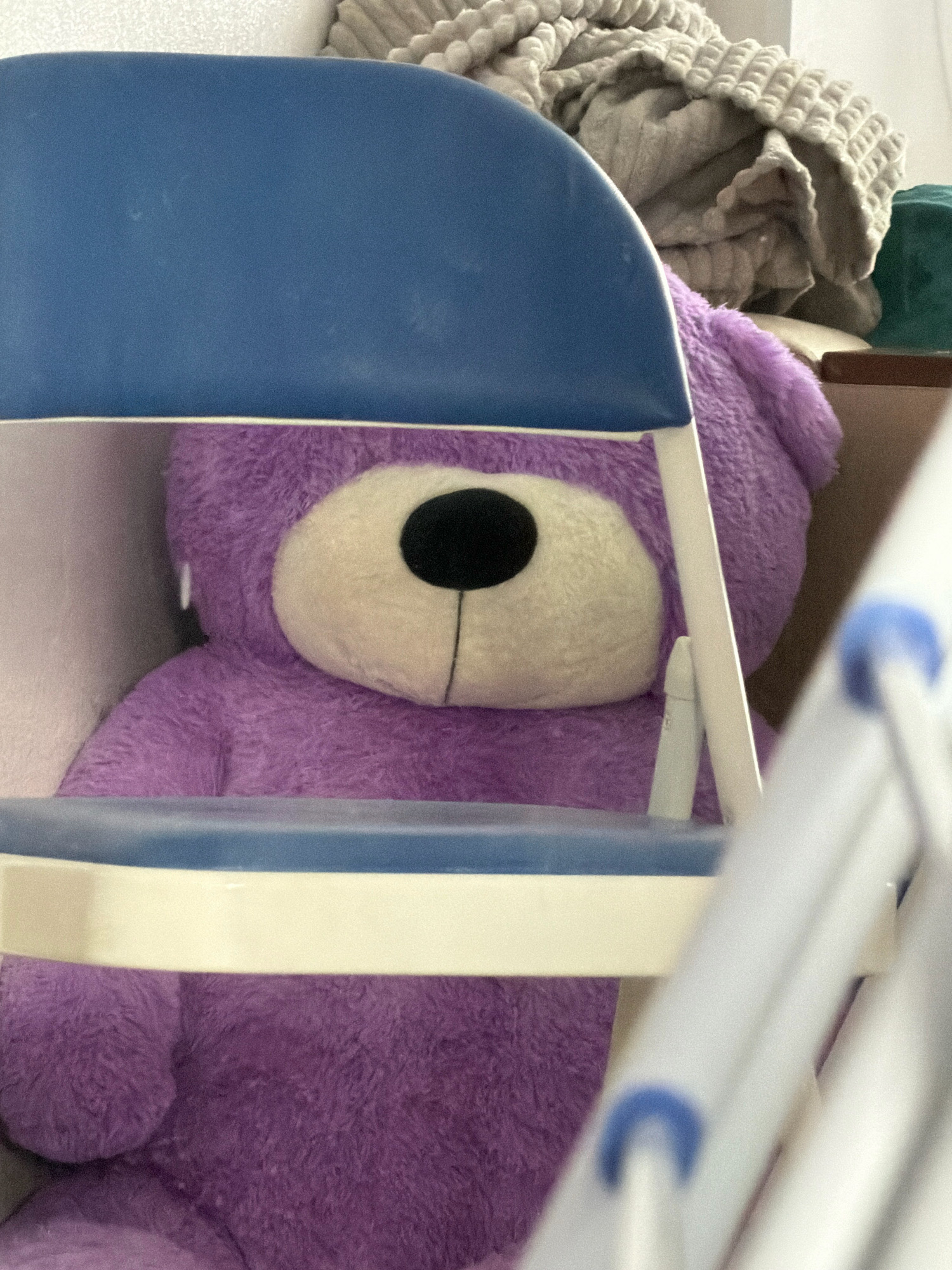 An indoor scene features a large, fluffy purple teddy bear sitting behind a blue and white chair. The teddy bear's face, with its cream-colored muzzle and black nose, is partially visible. In the background, a pile of beige, textured blankets is stacked on a shelf. The environment appears to be a domestic setting, possibly a bedroom or living room, and there are no people present. The image is a close-up shot, focusing on the teddy bear and the chair, with other elements blurred in the background. There is no discernible information about the location within the city of Khabarovsk, Russia.
