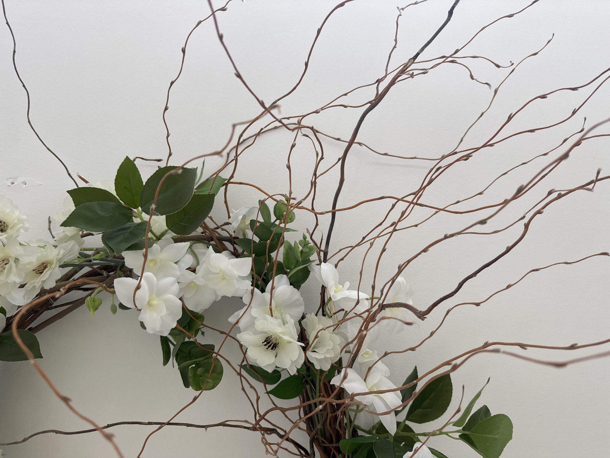 The image shows a floral wreath, predominantly composed of white flowers and green leaves, intertwined with thin, brown branches. The background is a plain white wall. There is no indication of the city of Chaikovsky, Russia, or any other specific location. There are no people or actions depicted.