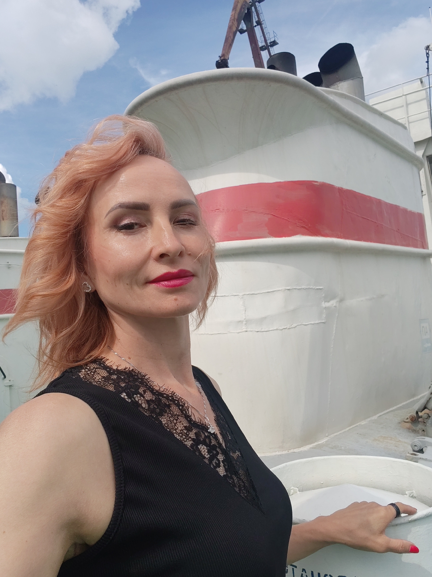 A medium shot captures a woman with light pink hair, wearing a black sleeveless top with a lace neckline and a silver necklace. She is posing for a selfie outdoors, with a large white and red structure, possibly part of a ship or industrial equipment, in the background. The sky above is partly cloudy. The woman is the primary subject, and the environment suggests an outdoor, possibly maritime or industrial, setting. The image does not provide any specific clues about its location in Selivanovo, Russia.