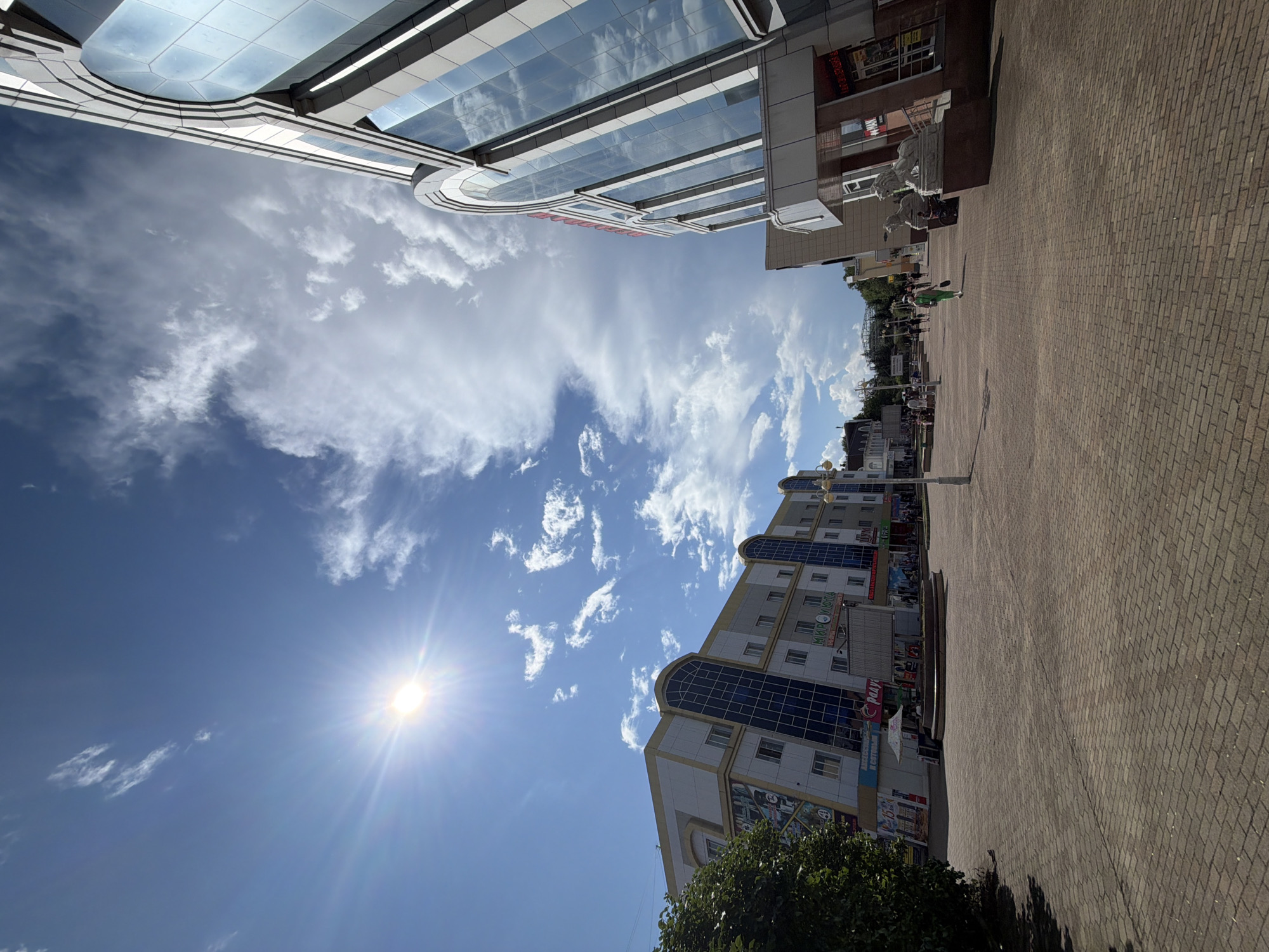 The image depicts a sun-drenched pedestrian area in Birobidzhan, Russia, featuring modern architecture and urban elements. The sky is a bright blue with scattered white clouds, and the sun is prominently visible, casting strong light and lens flares.
On the left side of the frame, a multi-story building with a beige and grey facade and blue-tinted windows stands tall. Its design includes arched window sections and signage, one of which reads "МИР УЮТА" (MIR UYUTA, meaning "World of Comfort"). A large tree with green foliage is situated to the left of this building.
On the right side, a contemporary building with a curved, glass facade is visible. This building also features arched window designs, reflecting the sky and surrounding environment. The ground level of this building has a more traditional facade with stone elements and entrances. Two lion statues are positioned in front of this building's entrance.
The pedestrian area is paved with brick-like tiles laid in a herringbone pattern. Several people are walking in the distance, suggesting a public urban space. In the middle ground, there's a circular fountain structure, and a small market stall with an umbrella is set up near it. Streetlights are present, and in the far distance, some taller structures and possibly a roller coaster or similar amusement ride can be discerned. The overall impression is of a lively city center on a clear, sunny day.