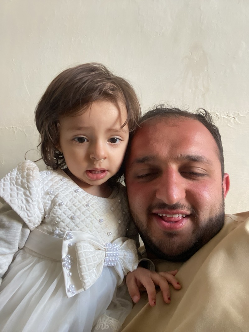 A close-up shot shows a man and a young girl posing closely together. The girl wears a white dress adorned with pearls and a large bow. The man, possibly her father, has dark hair, a beard, and is wearing a tan shirt. The background is a plain, light-colored wall. There is no verifiable information about the location being in Kandahar, Afghanistan.