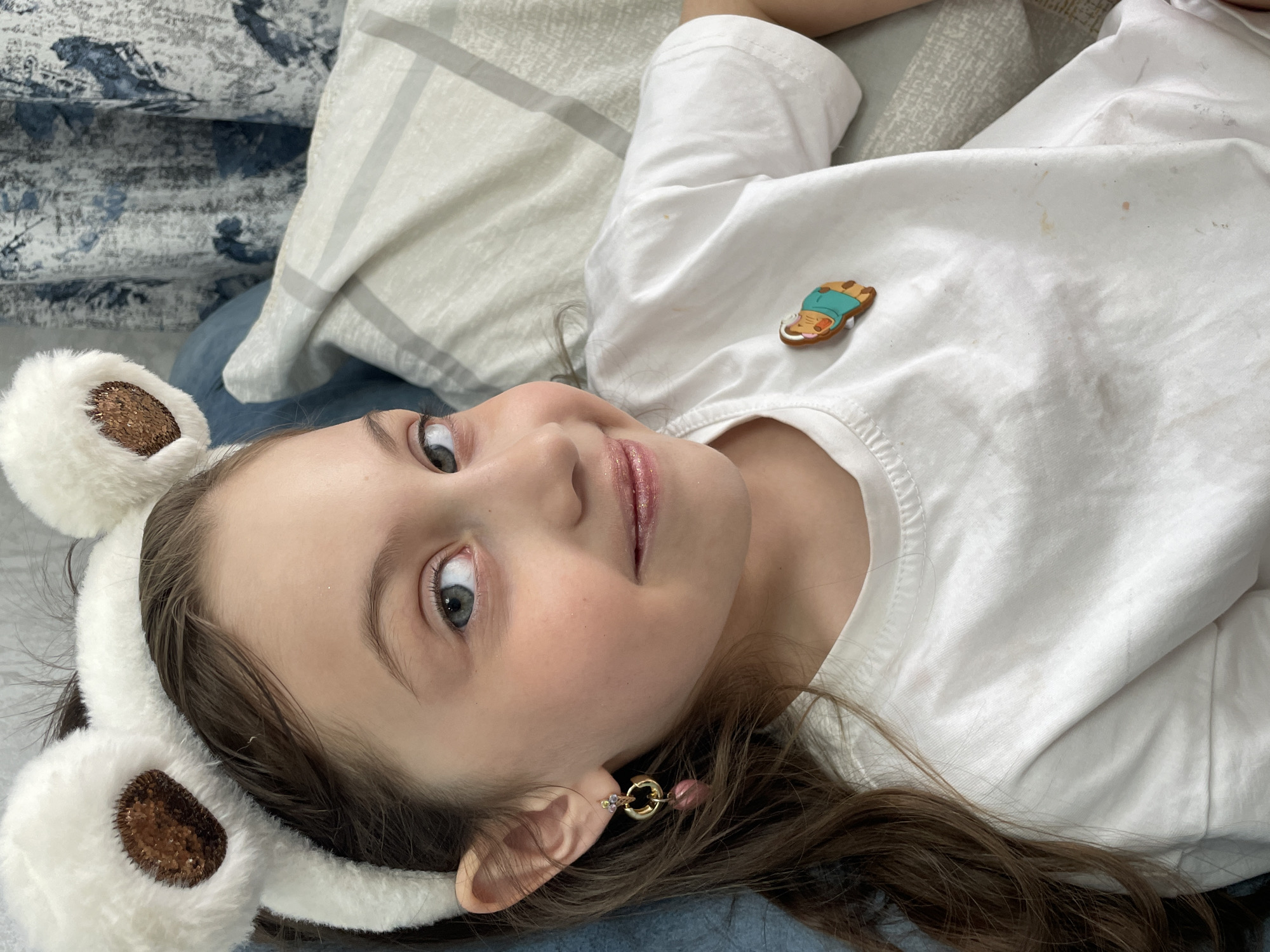 A close-up, eye-level portrait shows a young girl lying down, looking directly at the viewer with a slight smile. The image is taken indoors, likely in Izhevsk, Russia, depicting a casual moment of relaxation.
The subject is a child with light skin, blue-grey eyes, and long brown hair. She is wearing a white long-sleeved t-shirt, which has a few faint smudges or stains. On her head, she wears a white, furry headband adorned with two rounded ears that have shimmering brown, glittery centers, resembling a bear or animal. A small, decorative pin or brooch is attached to her t-shirt on her left side (viewer's right); it depicts a brown, cartoon-like animal, possibly a cat or bear, lying on its stomach and wearing a light blue top. In her right ear (viewer's left), she wears an earring featuring a gold-colored hoop from which hangs a small pink bead and a larger, possibly faceted, pink bead. Her lips appear to have a subtle shimmer or gloss.
The girl's expression is pleasant and serene, conveying a sense of contentment. The background consists of soft bedding or pillows in shades of white, grey, and blue, suggesting she is lying on a bed or sofa. The lighting appears natural and soft, indicative of daytime. There is no visible text in the image.