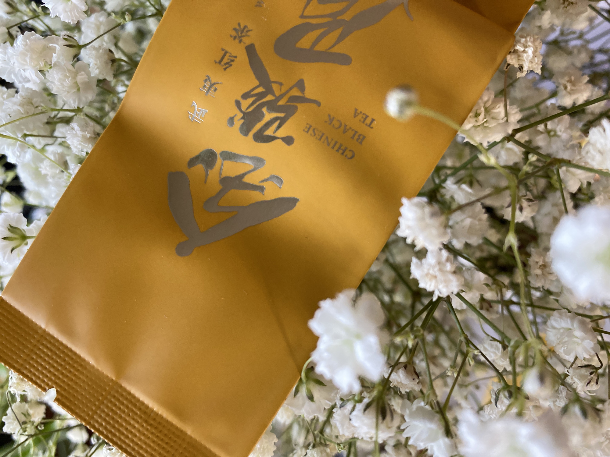 A close-up shot features a yellow packet of "Chinese Black Tea" nestled amidst a bed of white baby's breath flowers. The packet is gold colored and has Chinese characters and the English words "CHINESE BLACK TEA" printed on it in silver. The background is blurred due to the close proximity of the camera. No information is available about a location in the city of Chaikovsky, Russia.