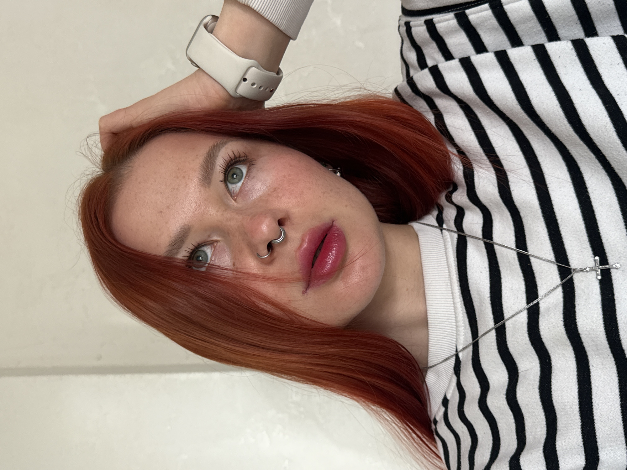 A young woman with fair skin, freckles, and red hair is posing for a selfie. She has a silver nose ring, light green eyes, and wears a white smartwatch. She is wearing a black and white striped shirt with a silver cross necklace. The background is a plain white wall. The location is not specified, and no evidence indicates it is Rostov-on-Don, Russia.