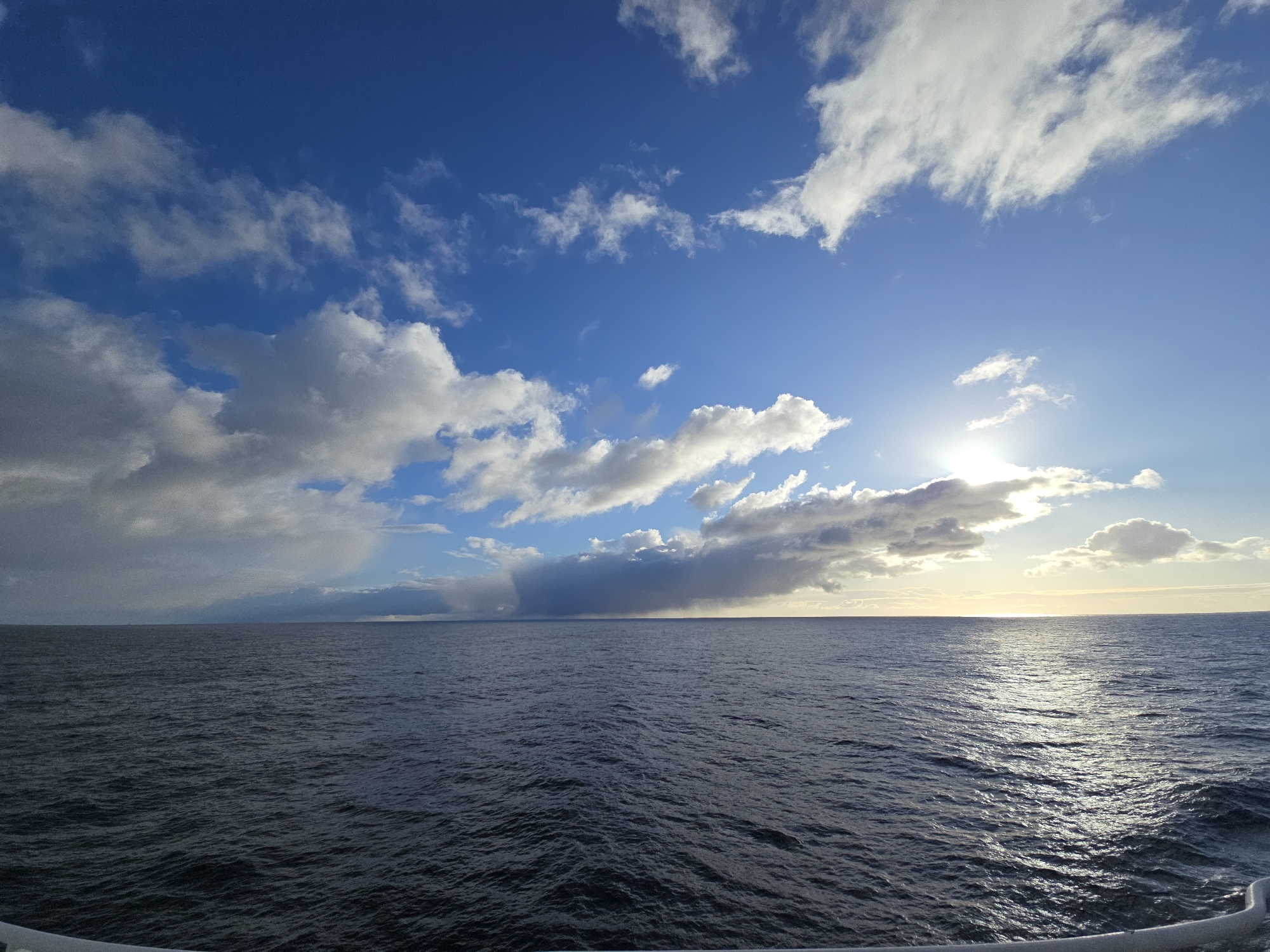 The image depicts a vast body of water, possibly an ocean or large sea, under a partly cloudy sky. The clouds vary in density, with some appearing light and fluffy while others are darker and more ominous, suggesting potential rain. The sun is visible, partially obscured by the clouds, casting a bright, diffused light across the water. The water itself is dark and rippled, indicating a slight breeze. The horizon appears distant and indistinct. The location is unknown. No people or interactions are visible in the frame. A small part of a white structure appears at the bottom of the frame, possibly part of a boat or ship.
