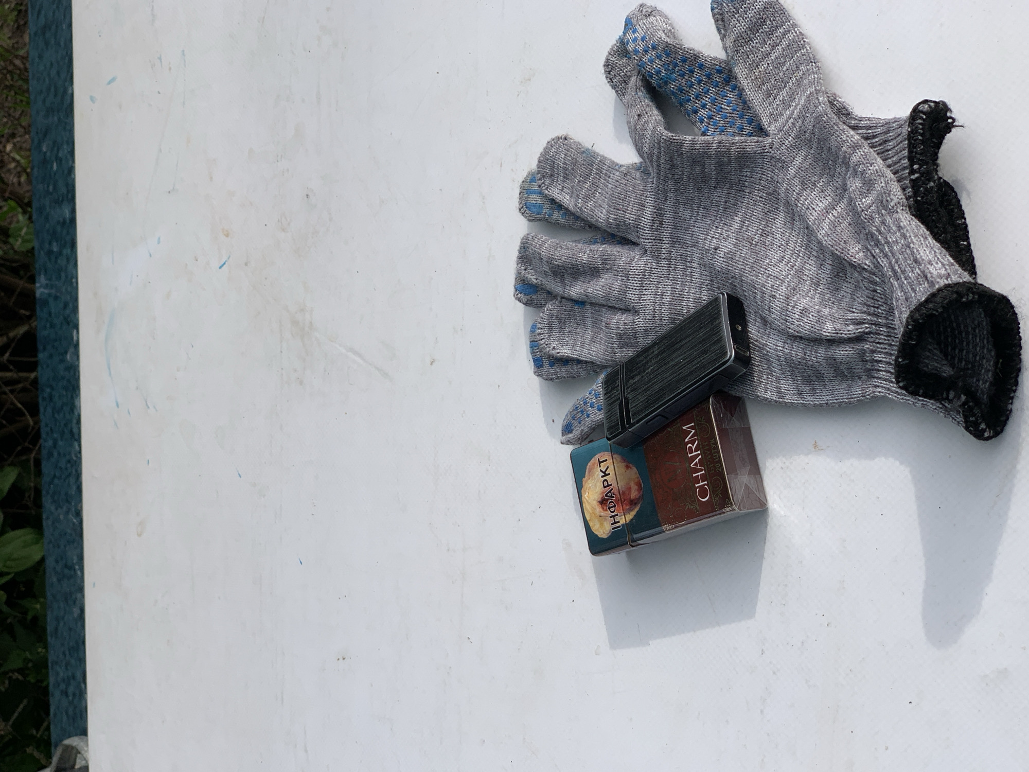The scene, located in Відзаўскі сельскі Савет, Belarus, displays a weathered, off-white surface with minor scuffs and faint blue markings. Three objects are present on the surface. A reddish-brown pack of "CHARM" cigarettes is visible, featuring a Cyrillic warning label reading "ІНФАРКТ" (INFARCT) alongside a graphic depicting a damaged heart. Adjacent to the cigarette pack, a black, textured electronic lighter rests. To the right, a single grey knitted work glove lies flat, characterized by blue rubberized dots on the palm and fingers and a black cuff. All objects cast shadows to their left, indicating a light source from the upper right. The background consists of out-of-focus green foliage positioned above a dark blue-green band.