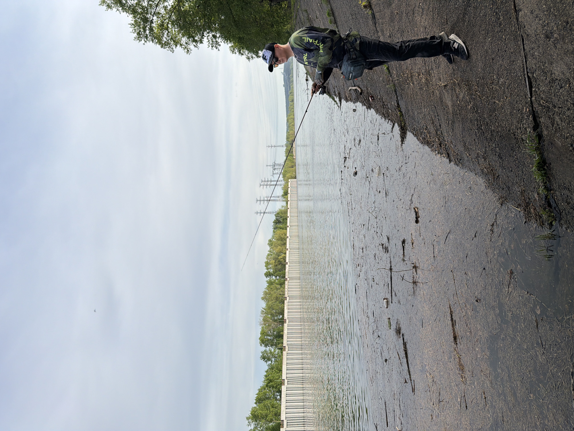 A man in a cap, green shirt, and black pants is fishing from a concrete embankment along a body of water. He is holding a fishing rod and looking down at the water. The embankment is wet and covered with debris. The body of water has a barrier extending across it, with trees visible behind the barrier. Power lines and towers are visible in the distance. The sky is overcast. The location is likely in the city of Chaikovsky, Russia, based on the presence of a barrier over a river and the known presence of such structures in the region.
