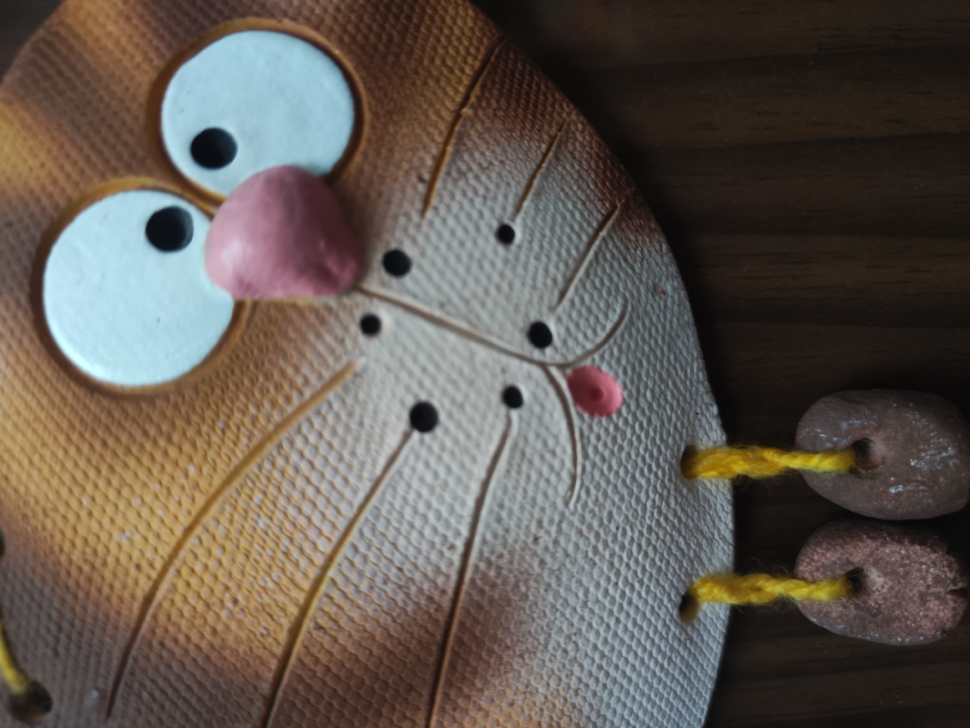 Close-up of a ceramic cat face decoration. The cat has large, white eyes with black pupils, and a pink nose. The face is textured, with brown stripes and black dots for whiskers. Yellow strings connect the face to two brown, oval-shaped objects on the right side. The background is dark wood. The image was not taken in Pushkin, Russia, as there is no indication of this in the image.
