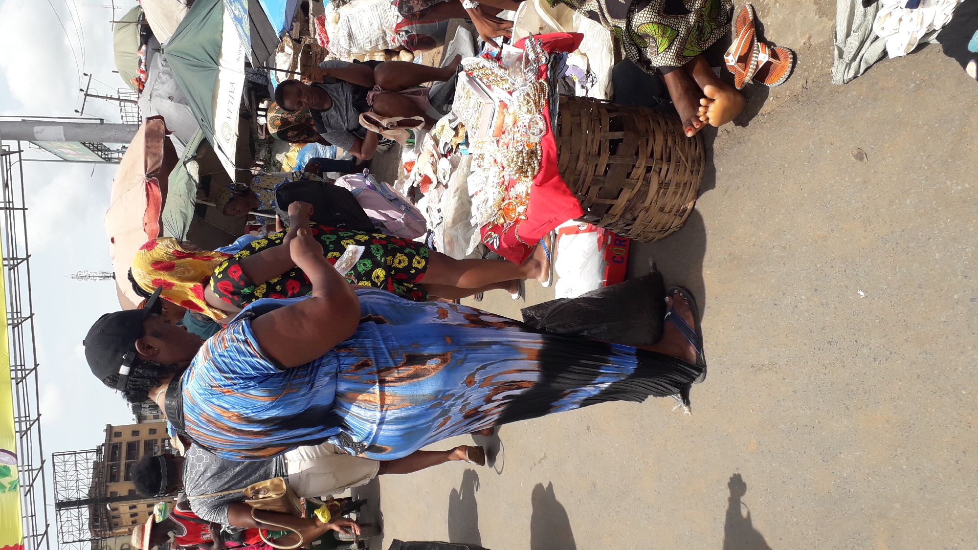 The image depicts a bustling scene in Onitsha, Nigeria, characterized by open-air market activities. Several individuals are visible, including women in colorful patterned clothing, engaging in commerce or browsing goods. A vendor sits surrounded by their wares, including beaded jewelry and fabric, displayed on tables and within woven baskets. The setting is a street or pedestrian area, partly shaded by umbrellas to offer protection from the sun. Buildings are visible in the background, suggesting an urban environment. The ground is paved and appears worn, reflecting the heavy foot traffic typical of such markets.