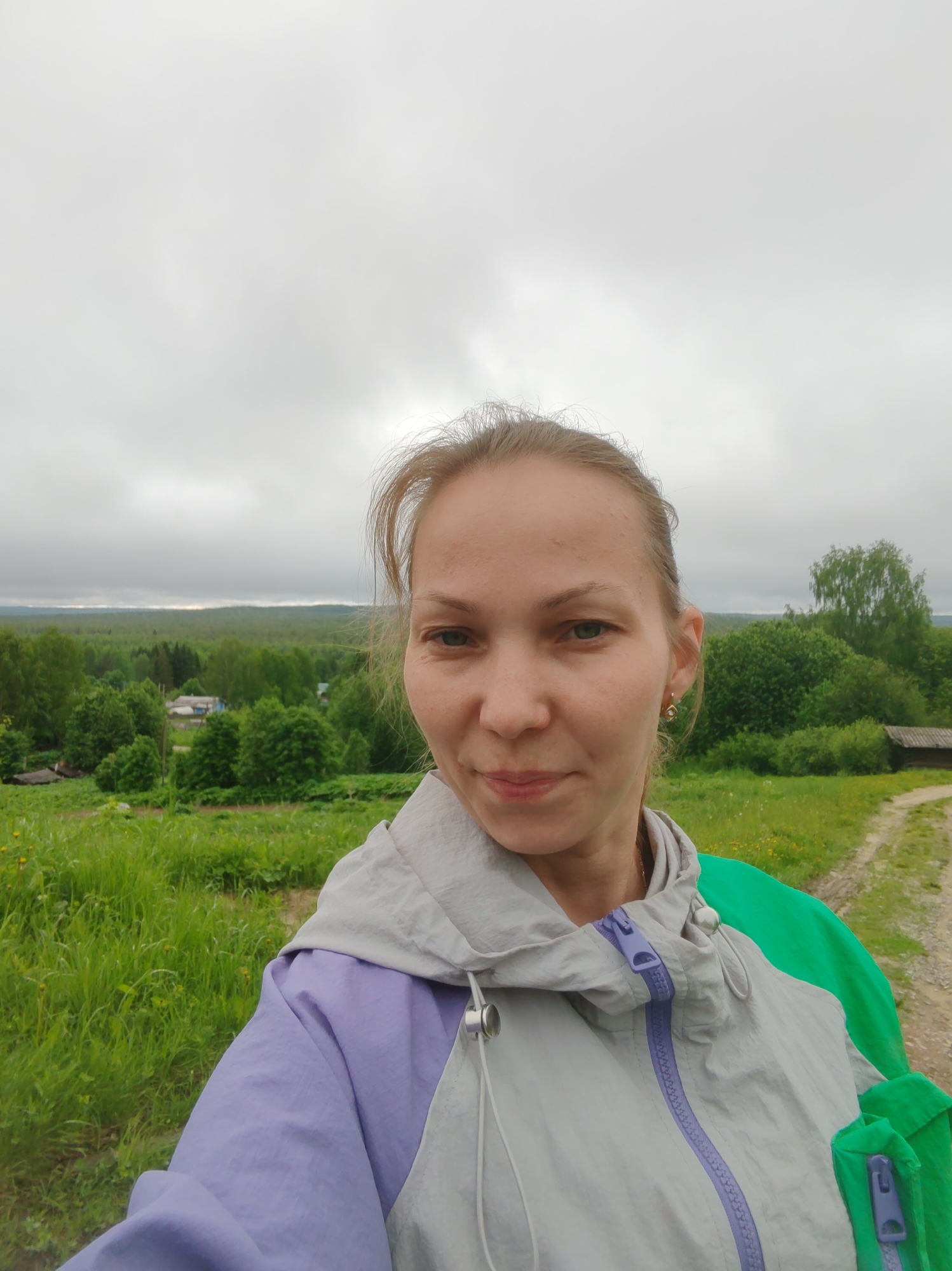 A woman stands in a grassy field in Чухлэм, Russia. She is wearing a grey, green, and lavender jacket. A forest is visible in the background under a cloudy sky. A dirt path runs through the field to the right of the woman.