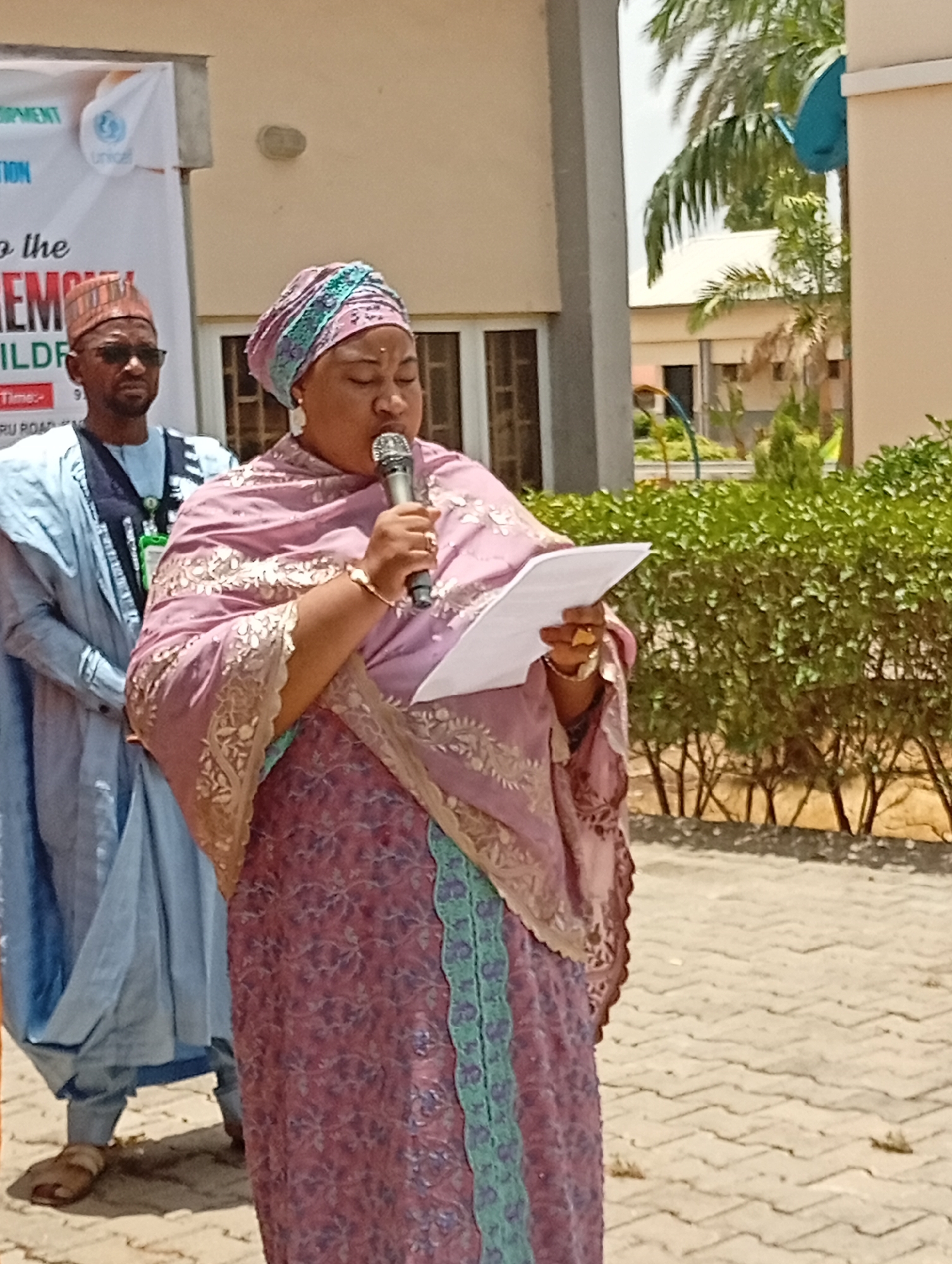 The image depicts an outdoor event taking place in Unguwar Sarki, Nigeria, under bright, sunny daylight conditions. In the foreground, a woman, appearing to be the main speaker, stands centrally, holding a silver microphone in her right hand and a sheet of paper in her left. She is dressed in a traditional, elaborately patterned pink and purple garment with gold embroidery, complemented by a matching headwrap featuring teal accents. Her expression is focused, suggesting she is either delivering a speech or reading from the paper. She wears visible gold jewelry, including large earrings, a bracelet, and rings.
Behind and slightly to her left, a man is partially visible, wearing a light blue traditional robe and cap, along with dark sunglasses. He stands attentively, observing the proceedings.
In the background, a banner is partially visible, indicating an organized event. Visible text fragments on the banner include "OPMENT" (likely part of "DEVELOPMENT"), the "UNICEF" logo and name, "TION," "to the," "REMONY" (likely "CEREMONY"), "CHILDRE" (likely "CHILDREN"), "TIME:", and "RU ROAD." This suggests a ceremony, possibly focused on children or development, with UNICEF as a key organizer.
The setting features a paved ground surface and low green bushes lining the background. Behind the subjects, a light-colored building is visible, and palm trees rise in the distance, indicating a warm, possibly tropical, environment. The strong shadows and clear sky confirm it is daytime.