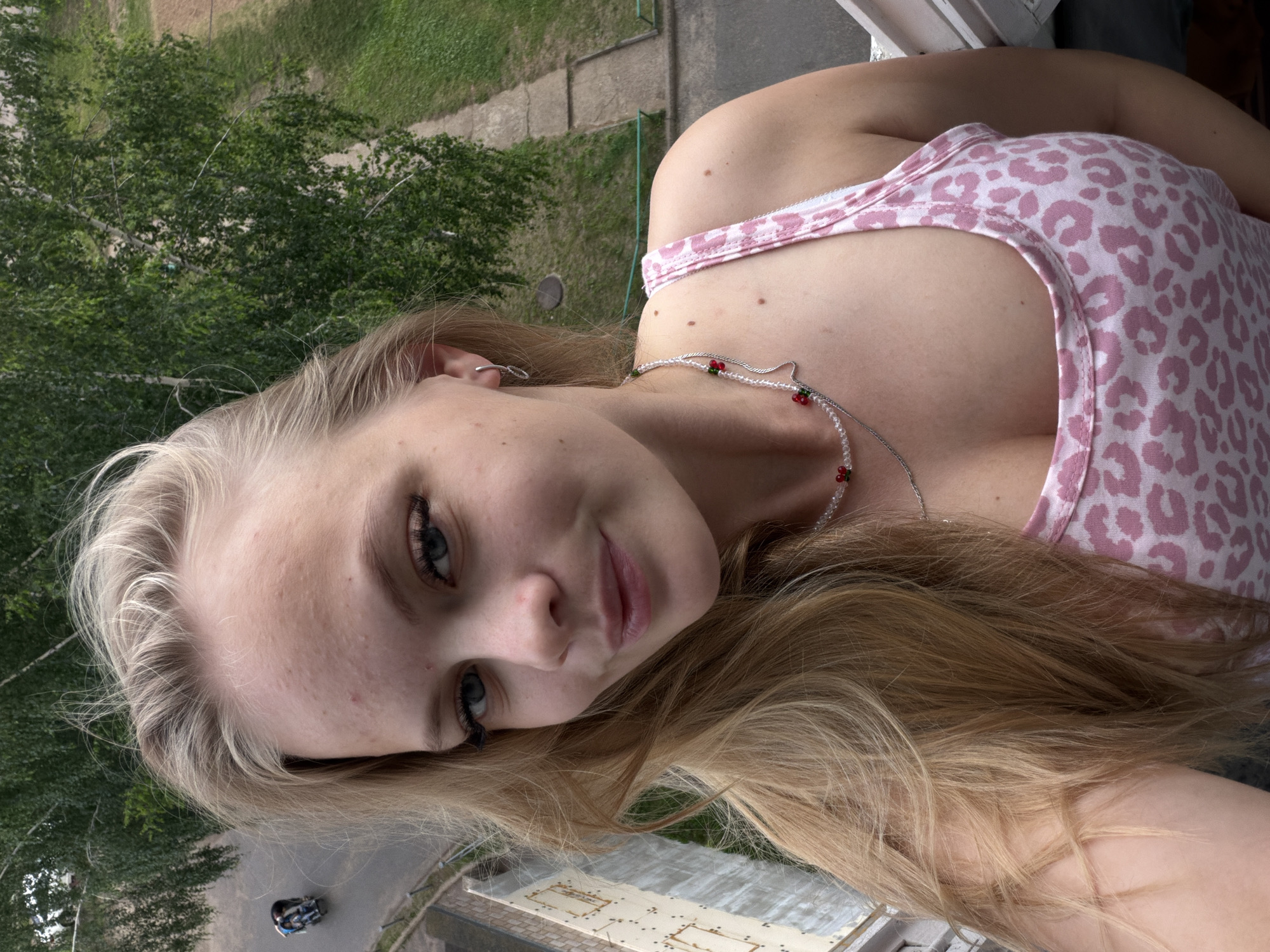 A young Caucasian female with blonde hair and blue eyes is taking a selfie outdoors. She is wearing a pink and white leopard print tank top and a necklace with clear beads and red accents. Her hair is partially obscuring her face and shoulder. In the background, there are trees, a sidewalk, and a building. A person on a scooter is visible on a road in the distance. The scene appears to be in a residential area of Chaikovsky, Russia.