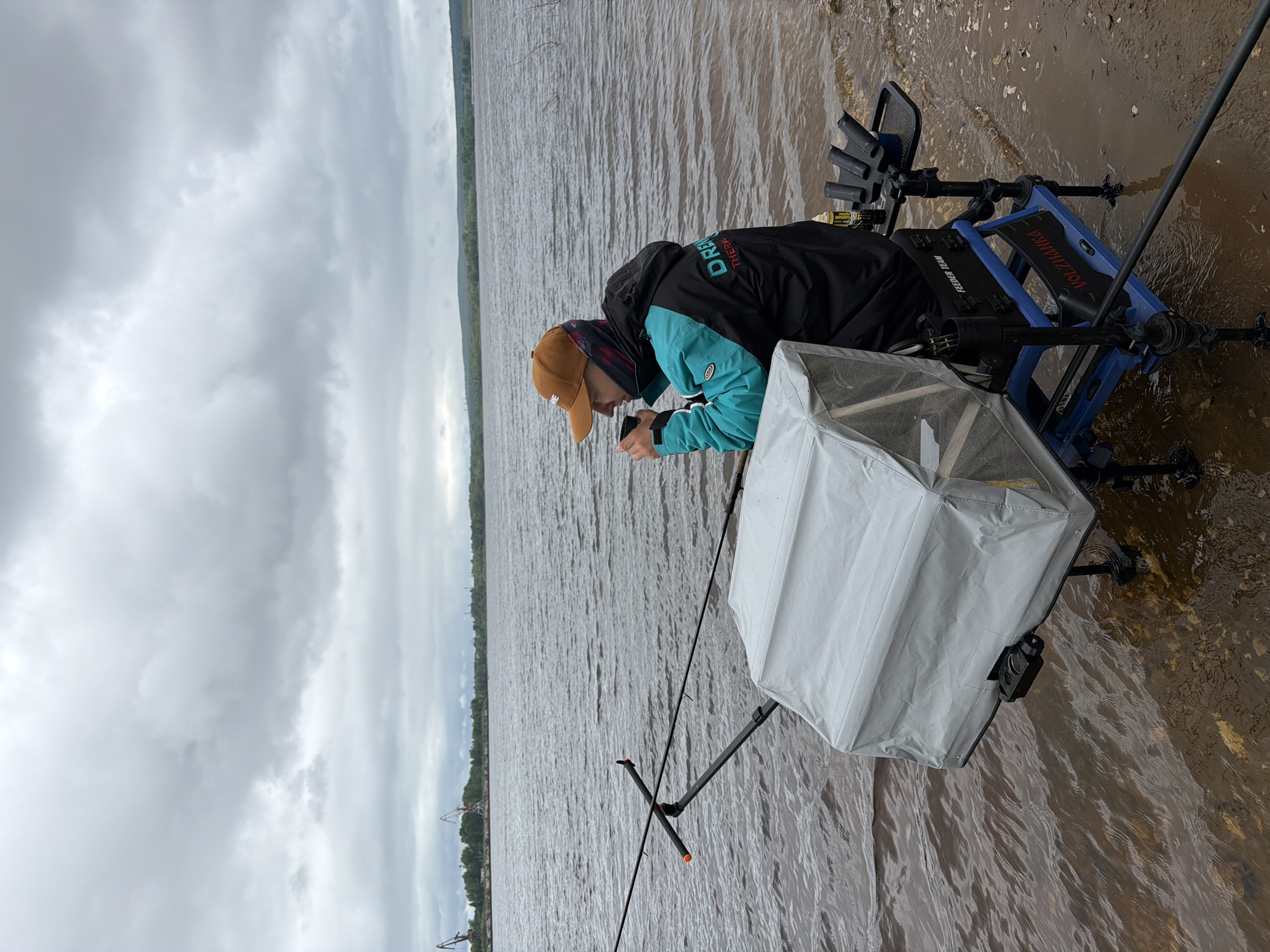 This image depicts a single individual engaged in feeder fishing on a muddy bank beside a large, murky body of water in Chaikovsky, Russia. The scene is set under a heavily overcast sky with thick grey clouds.
The individual, seen from the right side, is an adult male wearing a brown baseball cap, a patterned dark balaclava or neck gaiter, and a black and teal jacket with "DREEM" and "THERMAL" visible on the back. He is holding a dark-colored cup to his mouth, seemingly drinking.
He is seated on a specialized blue and black fishing platform or seat box, branded with "VOLZHANKA" and "FEEDER TEAM." The seat box has multiple adjustable legs, some of which are partially submerged in the shallow water near the bank. To his left, a large, rectangular light-grey object, possibly a covered bait station or keepnet, is attached to the seat box. Two fishing rods are set up; one extends horizontally to the left, resting on a rod holder attached to the seat. On the right side of the seat box, a black tray holds various fishing accessories, including several rod rests and a small bottle.
The body of water displays ripples and has a murky brown hue. The shoreline is muddy and shallow. In the distant background across the water, a treeline is visible, interspersed with industrial structures and several prominent port cranes, indicating an industrial area or river port.