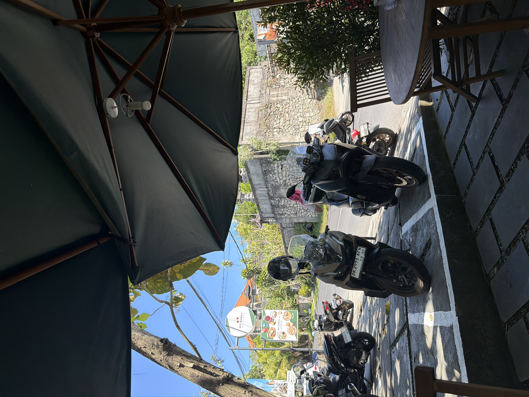 The image captures a street-level view of a cafe or restaurant seating area in Kuta Selatan, Indonesia. A large dark umbrella partially shades the foreground, revealing a wooden table and chairs. Several parked scooters and motorcycles are visible on the pavement, suggesting a common mode of transportation in the area. In the background, a stone wall, lush vegetation, and buildings indicate a tropical environment. A sign for "MY.BERRY" cafe is prominently displayed, advertising food and drinks. The sky is clear and blue, with the sun casting shadows. The overall scene conveys a relaxed, outdoor dining atmosphere in a bustling urban setting.