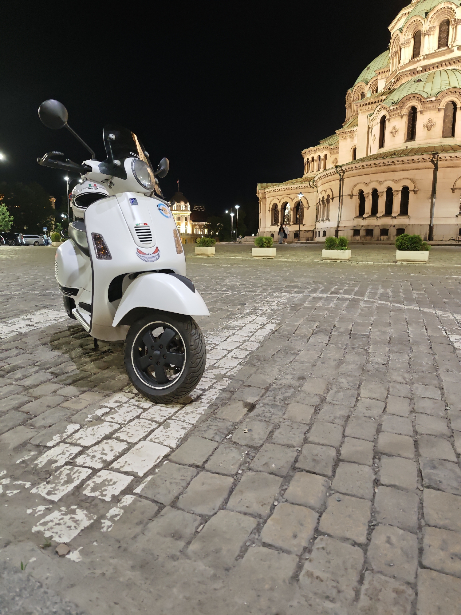 The image captures a stationary, white Vespa scooter parked on a paved square in Sofia, Bulgaria, at night. The Alexander Nevsky Cathedral dominates the background with its illuminated, multi-domed structure. The pavement consists of gray and white stone blocks. In the background, additional buildings, trees, and streetlights are visible. The scene appears quiet, with few people present.