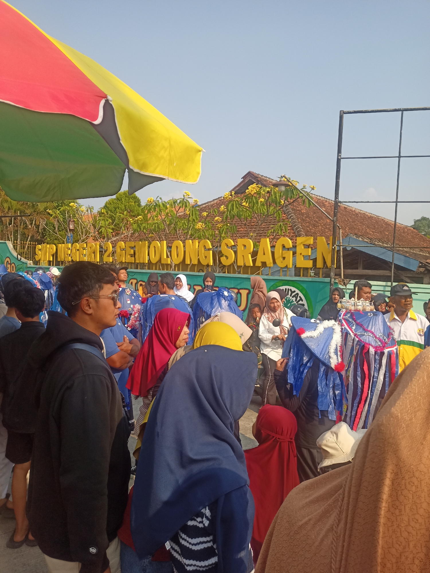 The image shows a street scene in Gemolong, Indonesia, with a group of people gathered in front of a building that reads "SMP NEGERI 2 GEMOLONG SRAGEN". The building appears to be a school, as indicated by the abbreviation "SMP" which stands for "Sekolah Menengah Pertama" (Junior High School). The people are diverse in age and gender, and many are wearing traditional Indonesian clothing, including headscarves. Some individuals are holding colorful decorations, possibly for a parade or festival. The environment is outdoors, with a clear blue sky and a large, colorful umbrella providing shade. The overall atmosphere seems festive and communal.