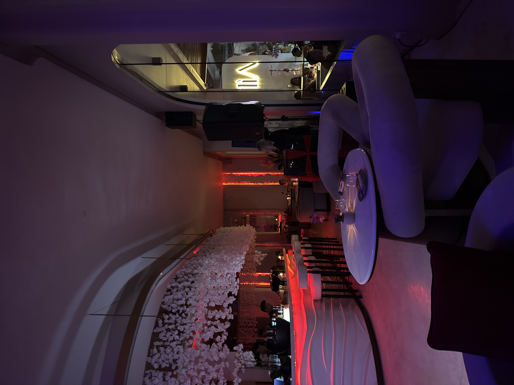 This media file depicts the interior of a dimly lit establishment, possibly a bar or lounge, with a modern and stylish design. The lighting is predominantly purple and red, creating a moody and intimate atmosphere.
In the foreground, a large, white, sculptural seating area dominates the right side of the frame, with a circular white table in front of it. Further back, a bar area is visible, illuminated by vibrant red LED lights. Several people appear to be present, some behind the bar, suggesting it is operational. There are also patrons seated at the bar and at tables further in the background, engaged in what seems to be a social gathering or enjoying drinks.
A prominent feature is a long, cascading arrangement of white artificial flowers that runs along a curved wall or partition, adding a touch of elegance and softness to the scene. In the background, a neon sign spelling "MVI" is illuminated against a wall, serving as a potential brand or decorative element.
The overall impression is that of a trendy evening venue, likely a bar or club, in a tropical location like North Kuta, Indonesia, given the context. The time of day is clearly evening or night, judging by the artificial lighting and indoor setting. There are no indications of weather or specific emotional cues, beyond the generally relaxed and social ambiance typical of such establishments.