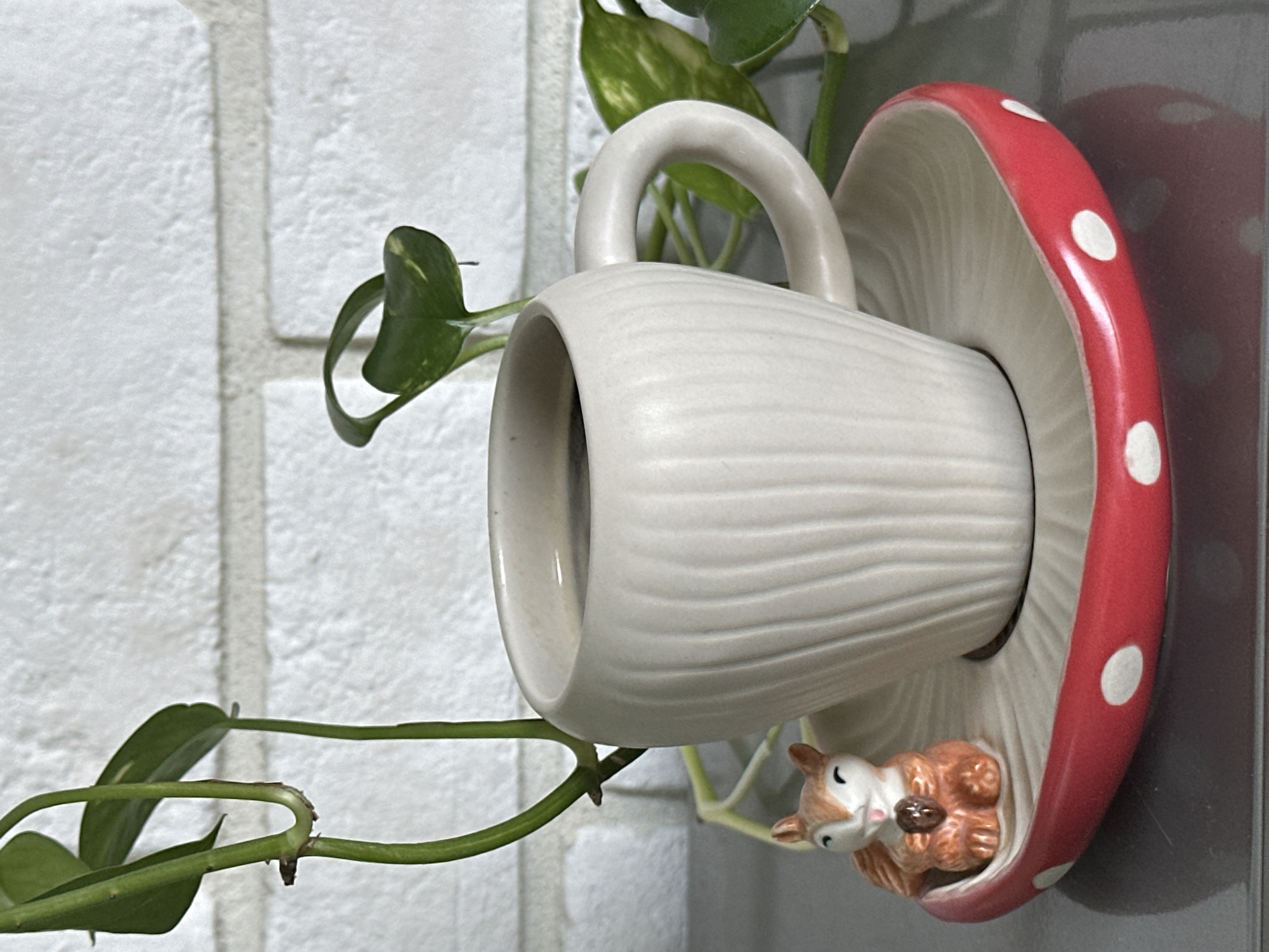 A close-up, eye-level shot captures a beige ceramic mug filled with dark liquid, resting on a white saucer with a red and white polka-dot rim. To the left of the mug, a small, orange ceramic squirrel figurine sits on the saucer, holding a brown nut. The saucer is shaped like a mushroom cap. Behind the mug, a green vine with leaves hangs down, with a white brick wall in the background. The setting appears to be indoors, with no indication of a specific location in Khimki, Russia.