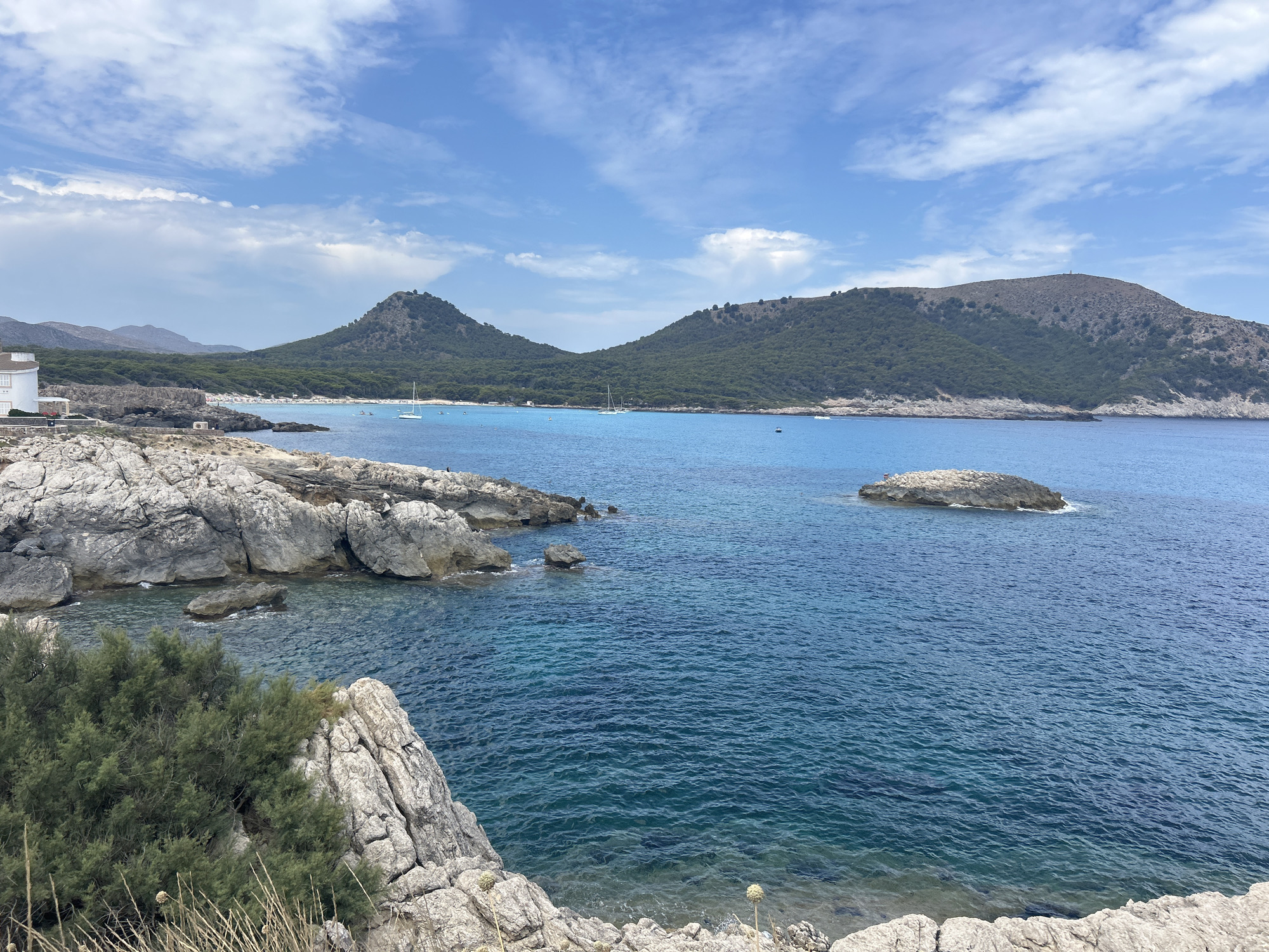 This is an eye-level, medium shot photograph of the coast of Cala Lliteres, Spain. The foreground is dominated by a rocky shoreline with a few sparse green bushes and dry grass. The rocks are light gray and appear rough and weathered. Beyond the rocks, the turquoise water of the Mediterranean Sea stretches out, with gentle ripples on its surface. In the middle ground, a small, isolated rock formation rises from the sea. Further out, several sailboats are visible, dotting the blue expanse. The background features two large, green, tree-covered hills that frame the bay, with a narrow strip of beach and some buildings visible between them. The sky above is a bright blue, with scattered white, wispy clouds. The lighting suggests a clear, sunny day, likely in the late morning or early afternoon. There are no people clearly discernible in the image, but the presence of sailboats and a beach suggests human activity in the area. The overall scene depicts a tranquil coastal landscape.