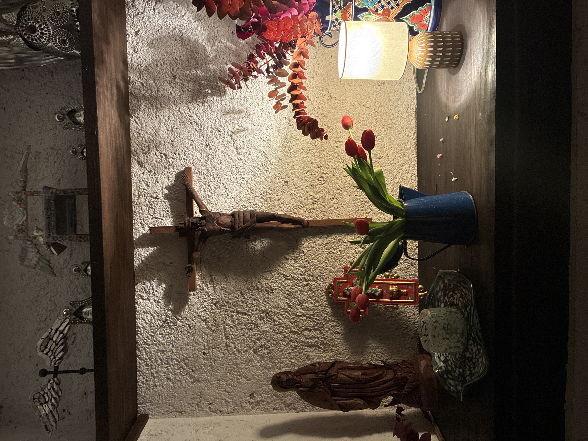 A scene presents a wall adorned with religious iconography in what appears to be a private residence, possibly located in the city of Cañada de Madero, Mexico, although this cannot be confirmed solely from the visual data. A crucifix with a dark, wooden figure of Christ dominates the center. To the right, a blue vase holds a bouquet of red tulips, with their vibrant color contrasting against the textured wall. Below the crucifix is a statue of a reclining figure, possibly depicting a saint or religious figure. A lamp, positioned near the top of the wall, casts a soft glow on the scene. There is also a decoration composed of red leaves and a ceramic plate. Adjacent to the vase of flowers is an ornamental glass piece. The wall itself has a rough, textured surface, and is surrounded by what appears to be wood trim. On the left side of the scene, there is an artifact made of metal, glass, and wood.