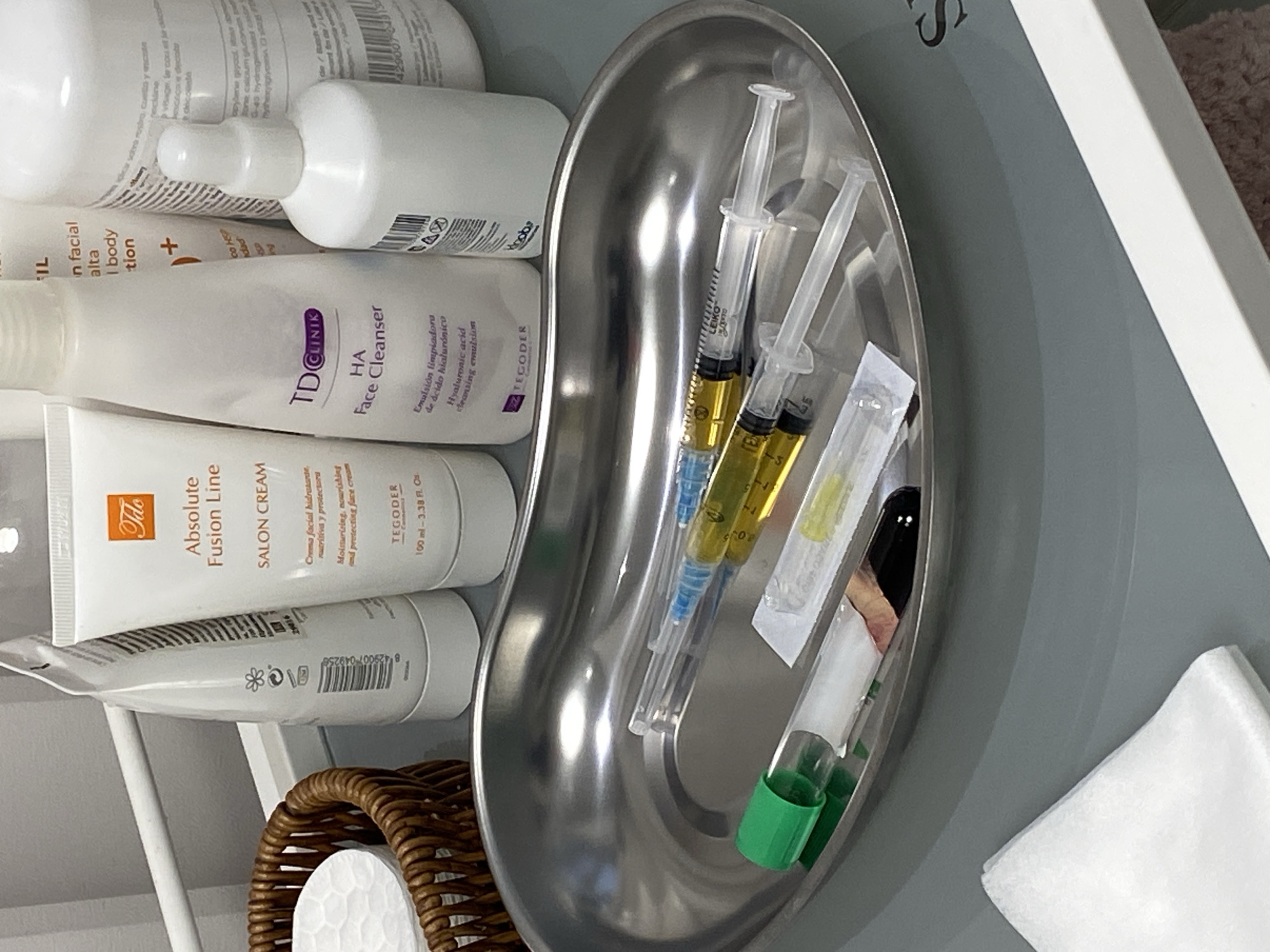 This eye-level, slightly tilted shot captures a collection of cosmetic products and medical supplies arranged on a grey surface, likely a trolley or countertop, in what appears to be a clinical, salon, or spa setting in Votkinsk, Russia.
On the left, several white bottles and tubes of skincare products are visible. Notable labels include "TD CLINIK HA Face Cleanser" (an "Emulsión limpiadora de ácido hialurónico" or Hyaluronic acid cleansing emulsion) and "Tito Absolute Fusion Line SALON CREAM" from "TEGODER," described as a "Crema facial hidratante, nutritiva y protectora" (Moisturizing, nourishing and protecting face cream) in a 100 ml - 3.38 Fl. Oz. container. Other products bear partial text such as "IL in facial alta I body tion +" and brand names like "Hoobs." Barcodes and small print, including numbers like "4290070" and "29016," are also discernible.
Dominating the right side of the image is a shiny metallic kidney dish. Inside it are two clear syringes filled with a yellowish liquid, possibly indicating platelet-rich plasma (PRP) or another serum, suggesting preparation for an injection-based cosmetic or medical procedure. Also in the dish are a smaller vial or test tube with a green cap and other wrapped sterile items, likely needles or swabs.
Partially visible at the bottom left is a brown wicker basket containing a white cotton pad or round. A white folded cloth or wipe rests on the grey surface to the far right. The lighting is bright and even, consistent with an indoor professional environment.