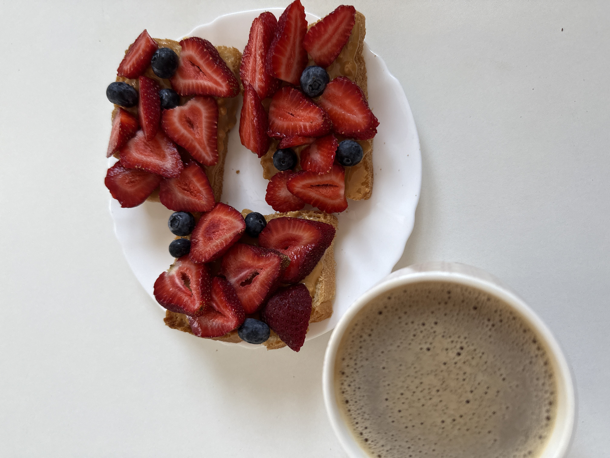 An overhead shot displays a breakfast arrangement on a stark white surface, likely a table or countertop, in an unknown location in Unknown, Unknown. Three pieces of toasted bread, each spread with a light brown, creamy substance consistent with nut butter, are positioned on a white ceramic plate with a subtly scalloped edge. Each toast piece is generously topped with an assortment of freshly sliced red strawberries and whole dark blue blueberries. The strawberries exhibit varying cuts, predominantly halves and slices, and are scattered amongst the darker blueberries. To the bottom-left of the frame, a white ceramic mug, partially cropped, contains a dark brown beverage topped with a light brown, frothy layer composed of small, uniform bubbles, characteristic of coffee with crema or a milk-based coffee. The background is uniformly white, devoid of additional objects or discernible features. No individuals are present in the image.