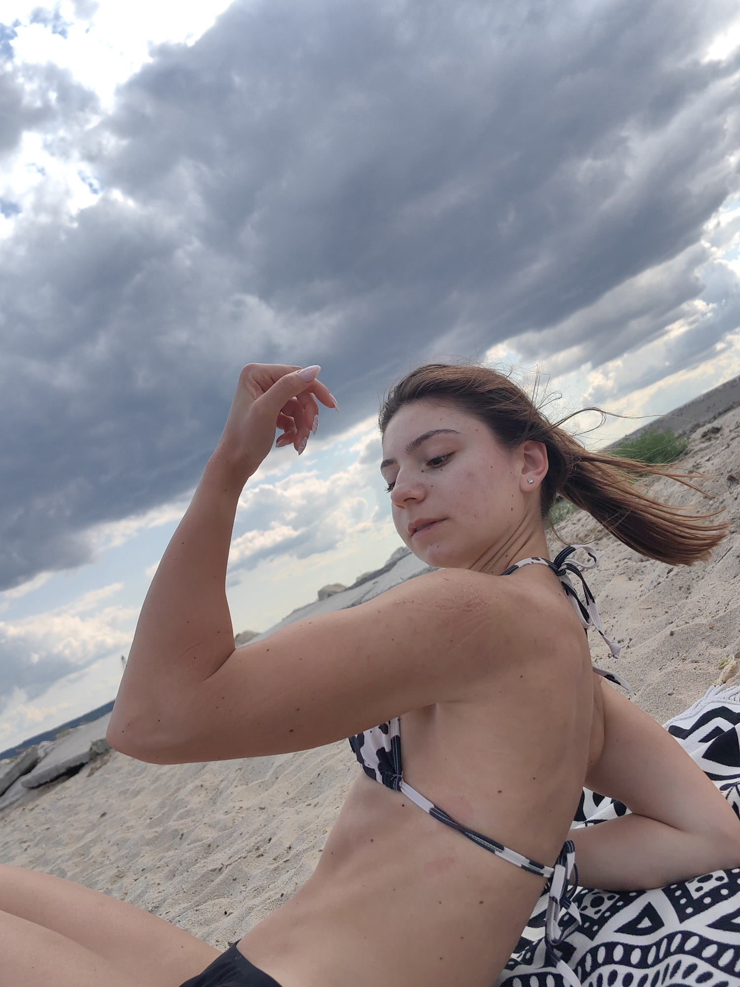 A young woman with brown hair pulled into a ponytail is sitting on a sandy beach, looking down and to her right. She is wearing a black and white patterned bikini. Her left arm is raised, with her hand resting near her head. Her right arm is bent, with her elbow resting on the sand. Behind her, the sand slopes upwards, and there are some large rocks and sparse vegetation. The sky is filled with dramatic, dark clouds, suggesting an overcast or stormy day. The light suggests it could be late afternoon or early evening. The scene appears to be a remote beach area, possibly near Varna, Bulgaria, given the provided location context.