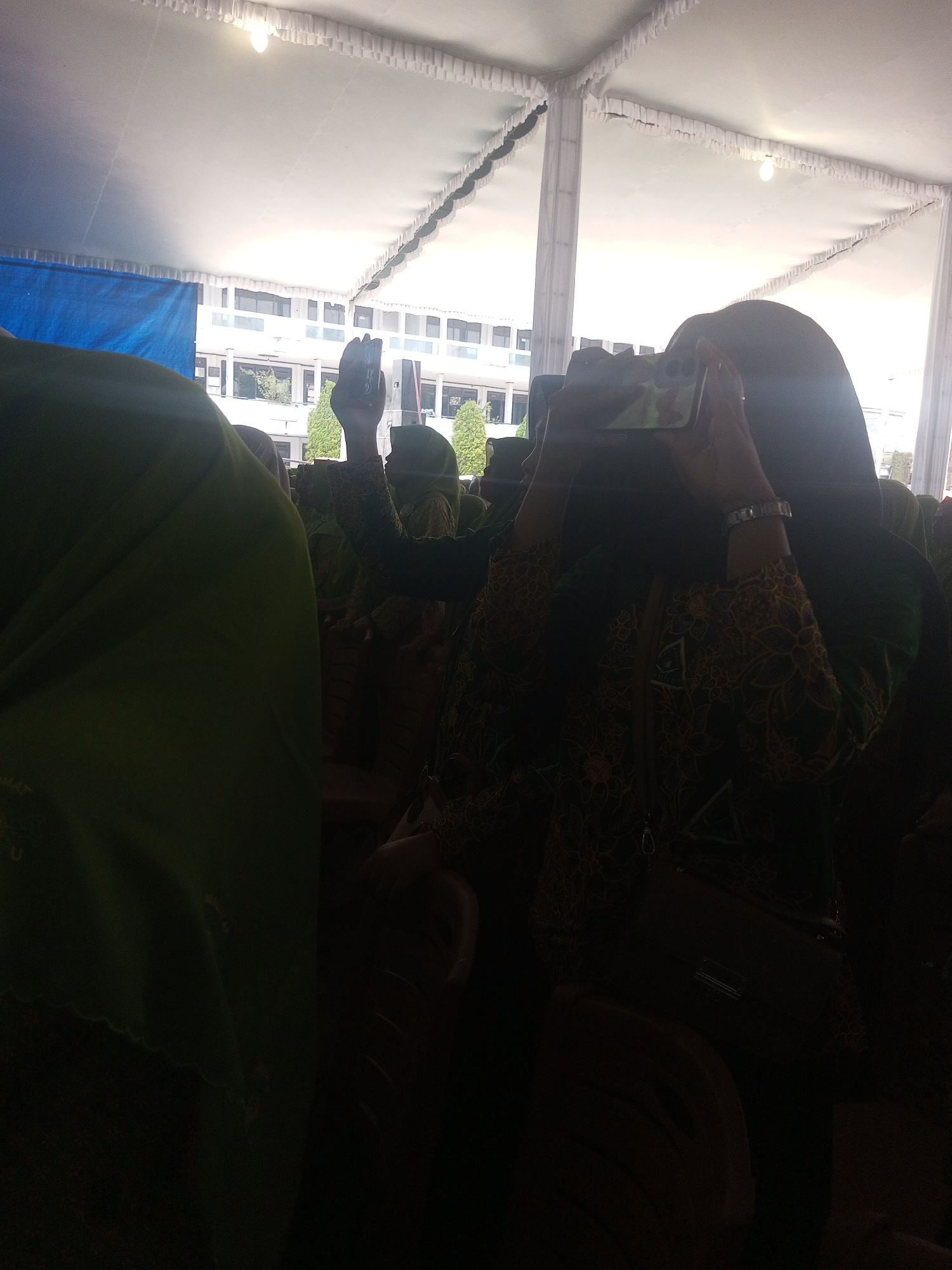 The image captures an outdoor event under a large white tent with a decorative fringe along the edges, likely in Klaseman, Indonesia. In the foreground, a person wearing a dark headscarf and a patterned blouse with green and gold motifs is holding up a mobile phone to record or photograph something. Their left hand is visible, and they are wearing a silver watch. To the left of this person, another individual is mostly obscured by a bright green fabric, possibly a garment or a banner. Behind them, a multi-story building with numerous windows is visible, suggesting the event might be taking place near an educational institution. Several other people in similar headscarves are also present in the background, some seated. The lighting is bright, with a single bulb visible in the tent ceiling, indicating it could be daytime or a well-lit evening event. The overall scene suggests a gathering or ceremony.