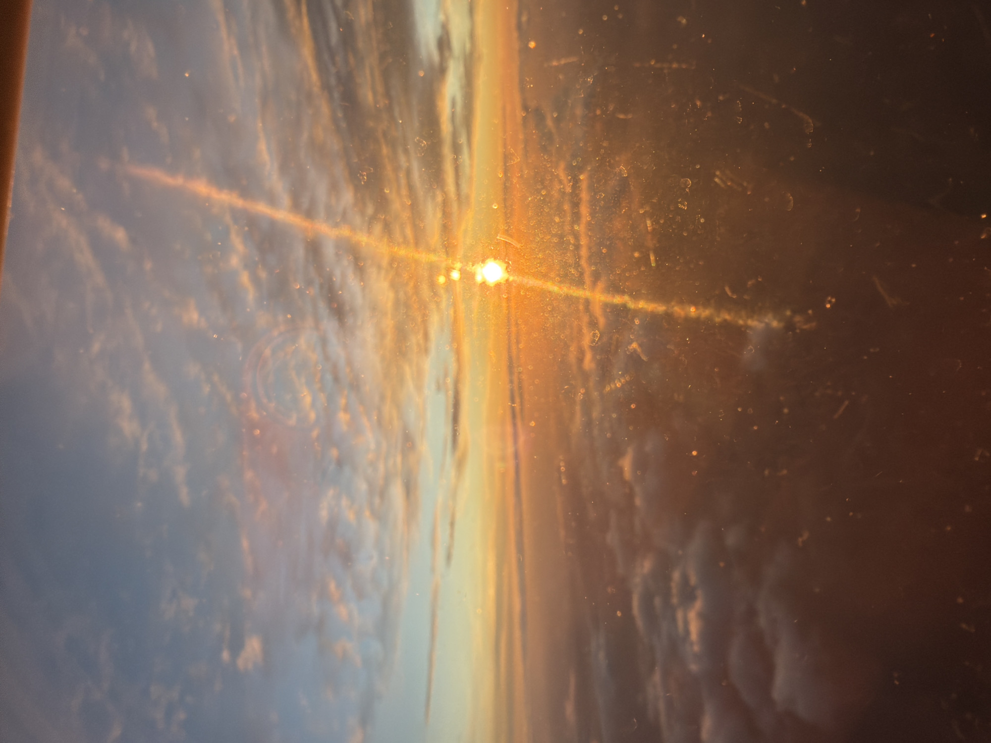 The image depicts a view from an airplane window, likely at dawn or dusk, showcasing a sunrise or sunset over a layer of clouds. The sun is a bright, luminous orb, with a strong beam of light extending diagonally upwards, creating a lens flare effect. The sky transitions from a pale blue at the top to warm oranges and yellows near the horizon. The clouds below are illuminated from above, appearing as soft, undulating masses of white and pinkish-orange. The window itself is visible, with numerous small specks of dust and smudges that refract the light, adding a textured quality to the image. There are no people, buildings, or other identifiable man-made structures in the scene, suggesting it was taken high above the ground. The location in Svidnya, Bulgaria, cannot be determined from the visual information provided in the image.