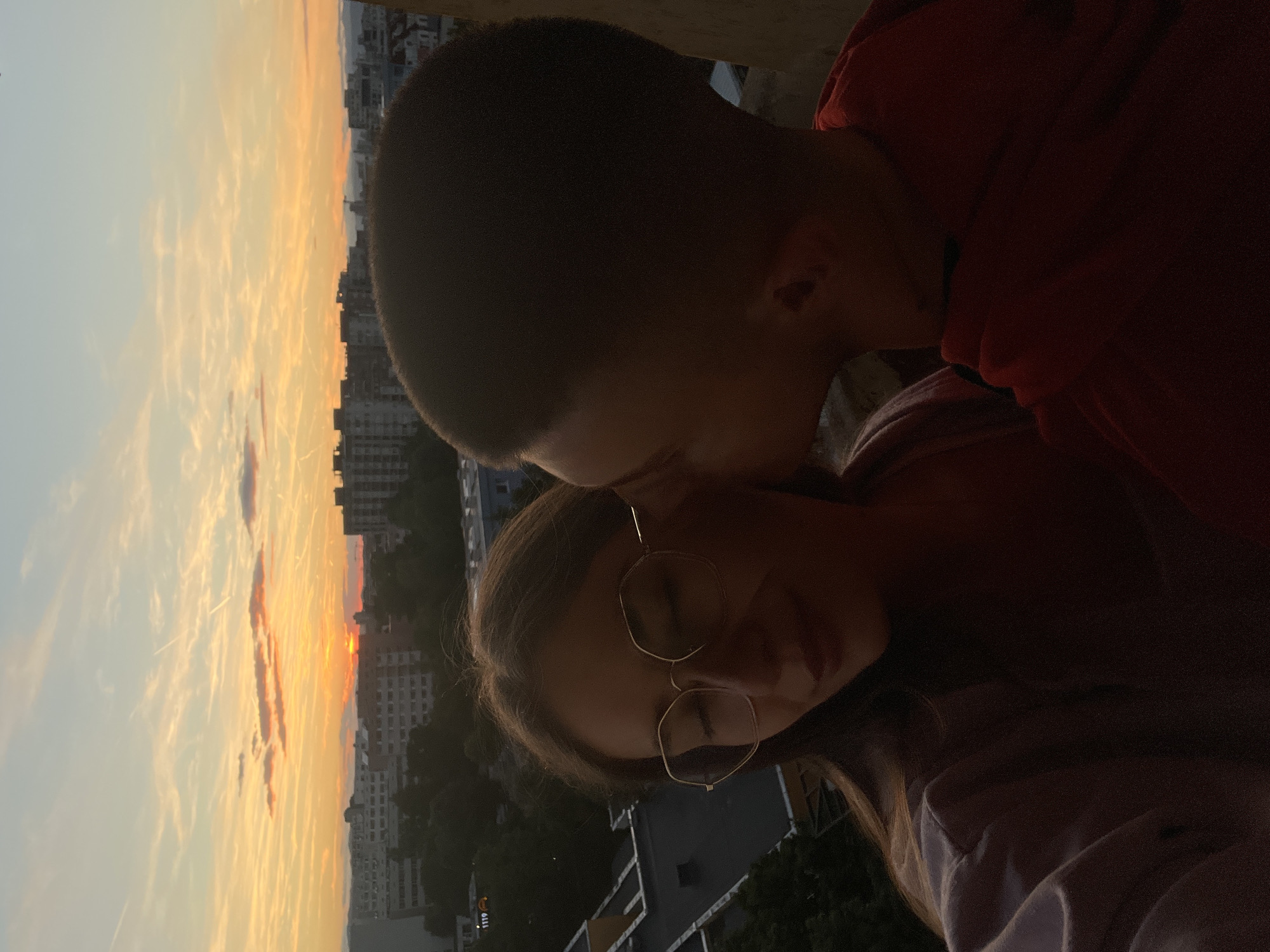 A couple is seen in a close-up shot during a sunset in an urban setting. The woman, on the left, is wearing large, gold-rimmed glasses and has her eyes closed. She is looking down, and her lips are slightly parted. She is wearing a light-colored top. The man, on the right, is leaning in and kissing her cheek. He has dark, short hair and is wearing a red hoodie. The background shows a city skyline with buildings and trees silhouetted against a colorful sunset sky. The sky is filled with streaks of orange, yellow, and pink from the setting sun, with some scattered clouds.