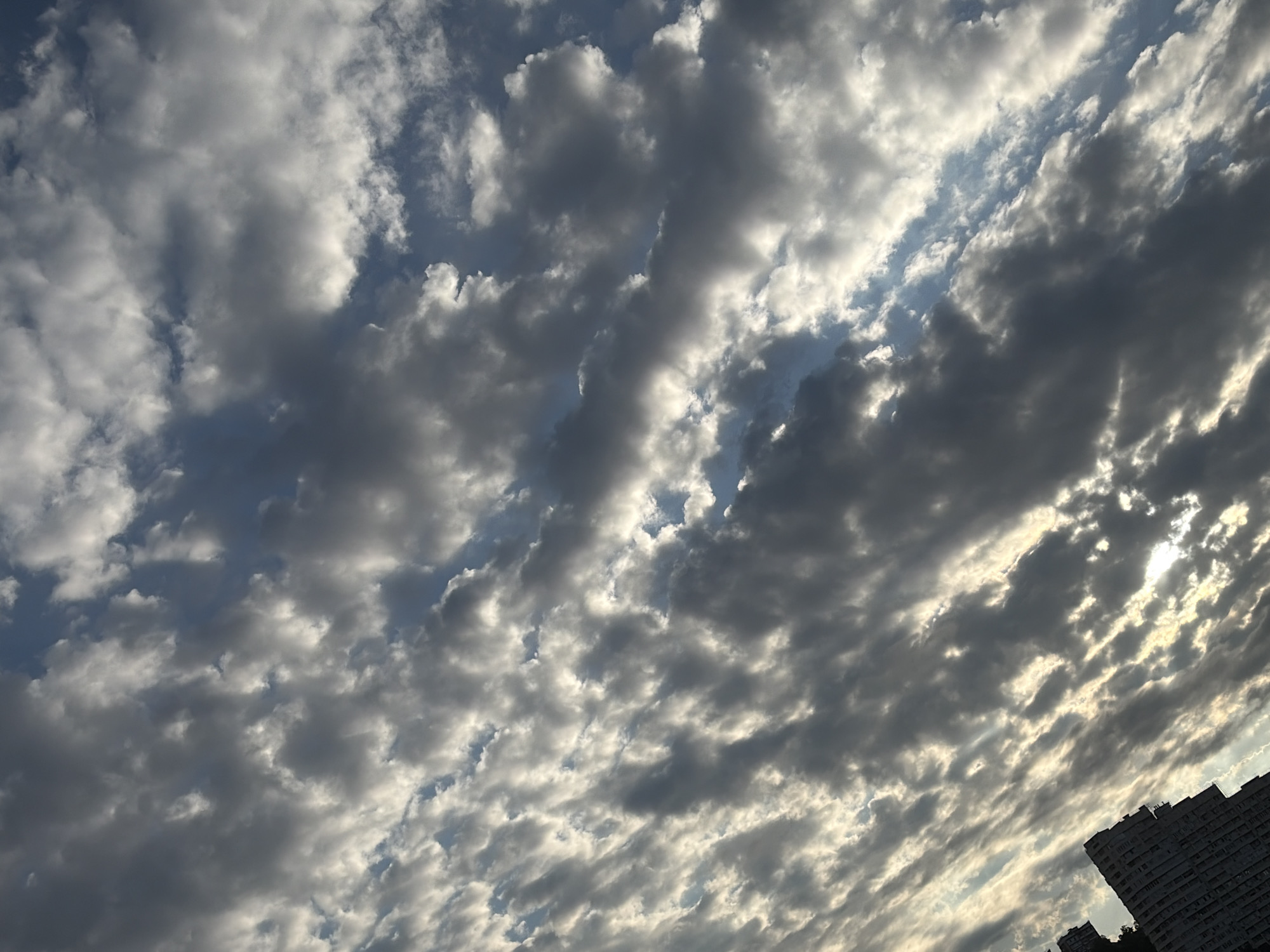 This is a high-angle, full shot of a cloudy sky with a building visible in the bottom right corner. The sky is filled with a mix of dark gray and white clouds, with patches of blue sky peeking through. The sun is visible through a gap in the clouds, casting a bright light. The building is a tall, multi-story structure with many windows, and appears to be an apartment building. The image was taken in Sochi, Russia.
