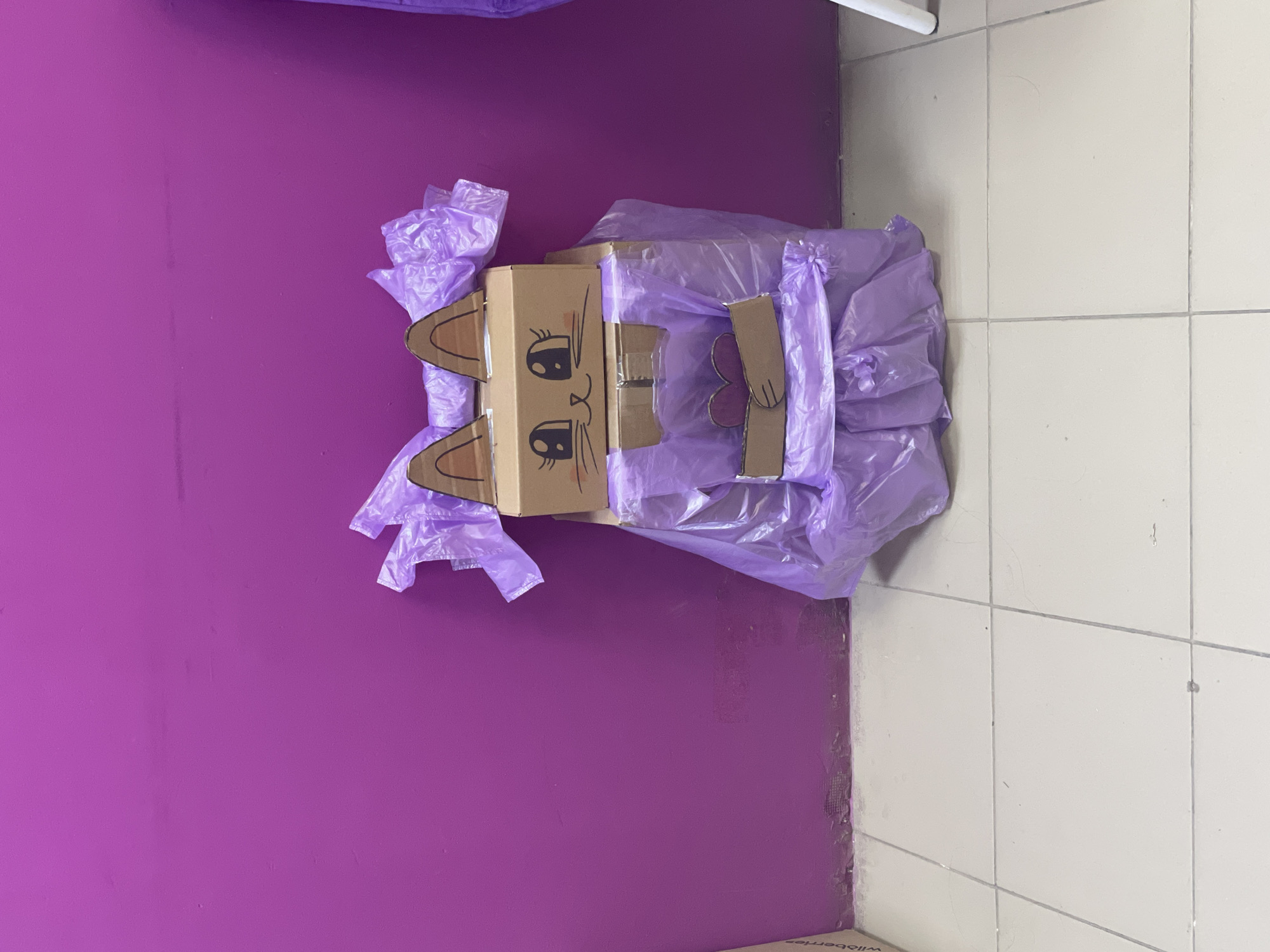 A cardboard box with a cat face drawn on it lies on a purple surface covered with several light purple plastic bags. The box has cardboard ears and cardboard arms holding a purple cardboard flower. The scene is inside a room, with a white-tiled floor partially visible on the right. The bottom right corner of the image has the word "wildberries". There is no indication of the location being Izhevsk, Russia.