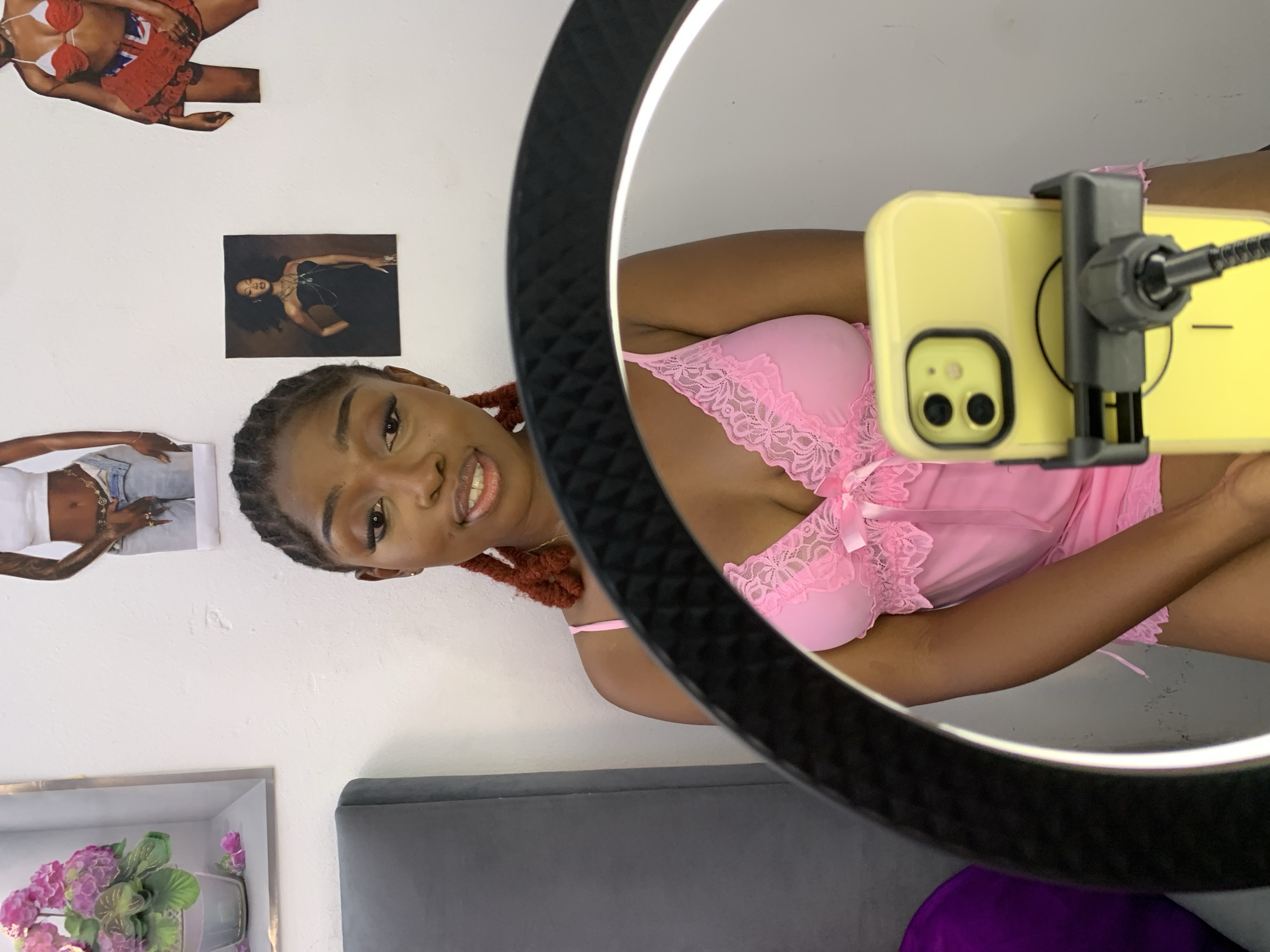 The image shows a young Black woman with braided hair, wearing a pink lace lingerie set, sitting in front of a ring light and a smartphone mounted on a tripod. She is looking towards the camera with a slight smile. In the background, there are several posters on the wall, including one with a floral arrangement and others featuring images of women in various poses. To the left, a gray headboard of a bed is visible. The overall setting appears to be a bedroom or a personal space, likely within a residential area in Lagos, Nigeria.