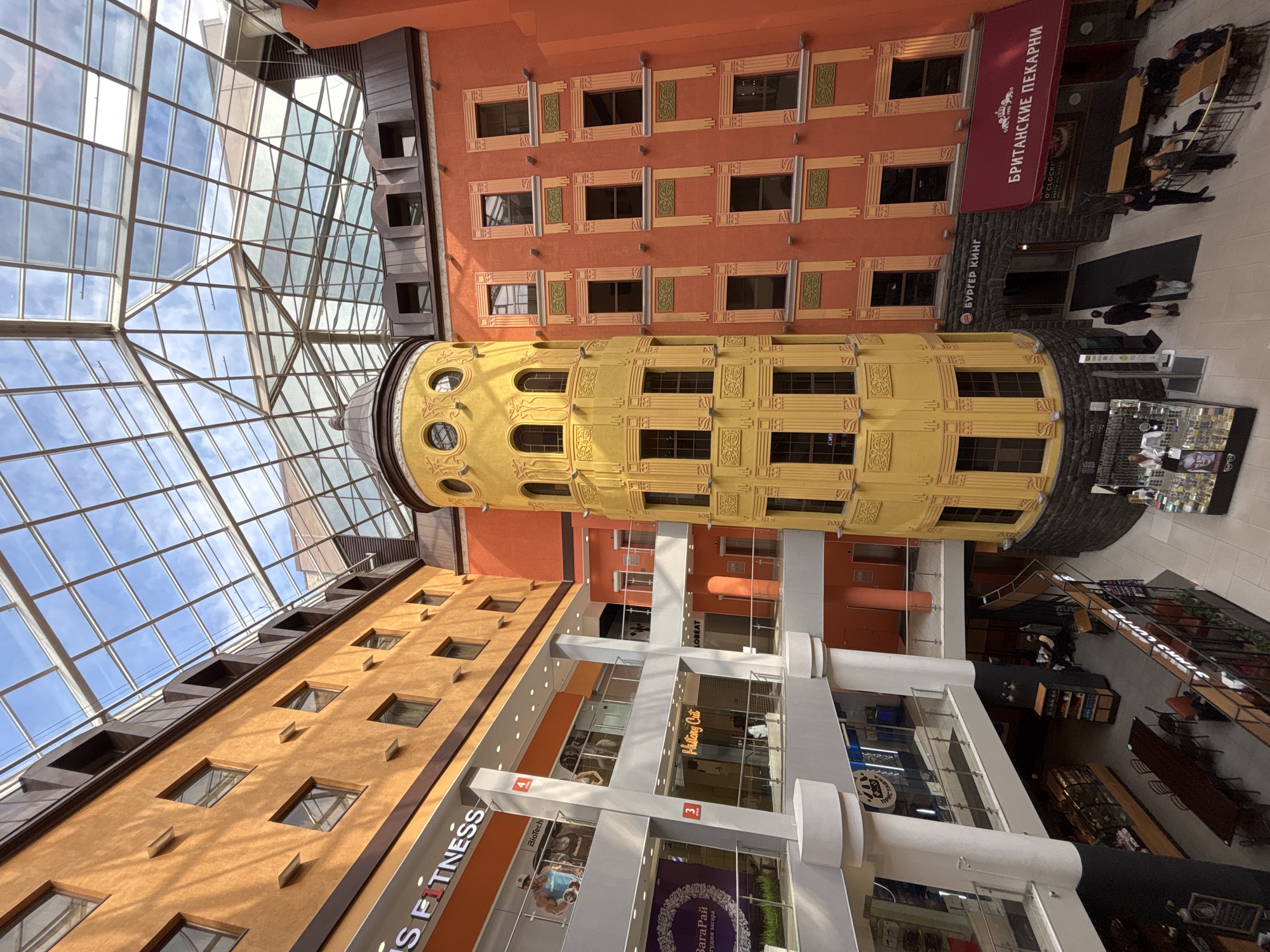 The image shows the interior of a shopping center in Saint Petersburg, Russia. It features a multi-level structure with several shops and restaurants, a yellow tower-like structure with windows, an orange building with decorative window frames, and a glass ceiling allowing natural light to illuminate the space.
On the ground level, there is a Burger King and a British Bakery (БРИТАНСКИЕ ПЕКАРНИ) with people walking by or sitting at tables. On the upper levels, there are other shops, including a fitness center (FITNESS), a tanning studio (ЗагаРай студия загара), and a hair salon (Hustong Cut). The architecture is a blend of styles, including ornate detailing and modern glass and steel. People are present, walking through the shopping center and occupying the seating areas. The image is taken from a low angle, looking upwards towards the glass ceiling.