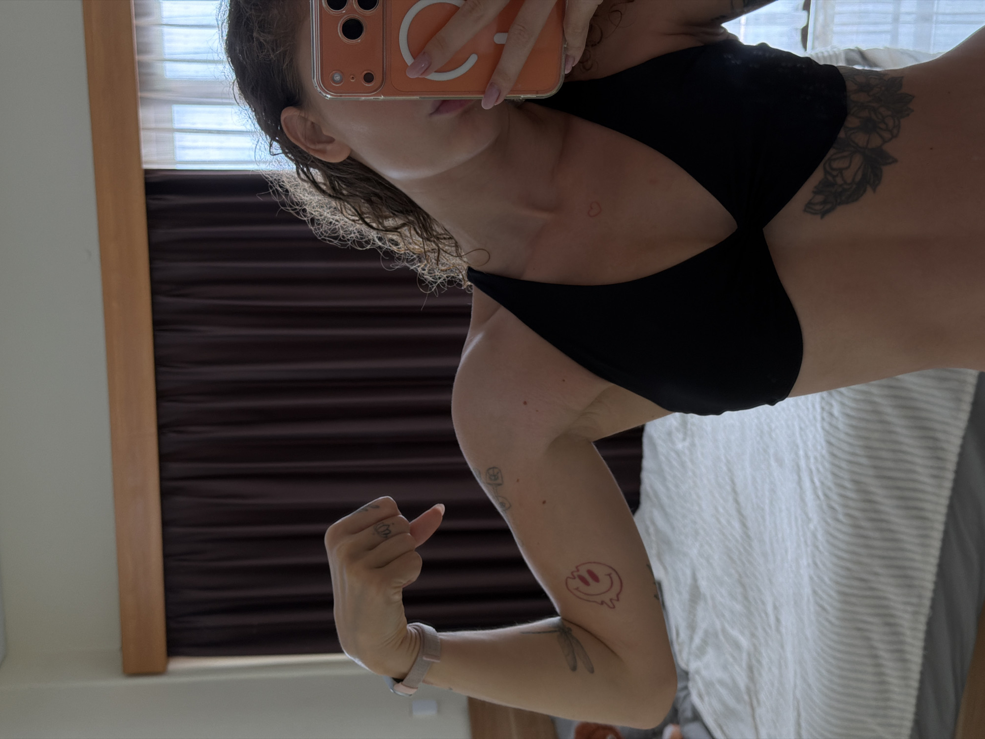 A woman is taking a mirror selfie in a bedroom. She is wearing a black sports bra. She has curly brown hair and several tattoos, including a dragonfly on her left arm, a melting smiley face on her left forearm, a small heart on her chest, and flowers on her right side. She is flexing her bicep on her left arm, and her right arm is holding a phone up to take the picture. The background shows a bed with white and grey striped bedding, dark brown curtains, and a wooden trim along the top of the window. The room appears to be in a home or hotel.