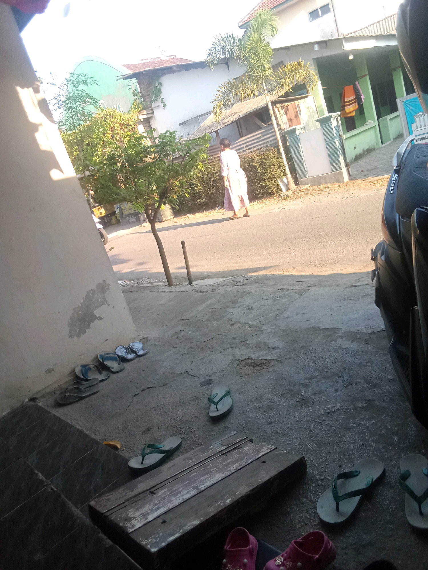 The image depicts a street scene in Jubung, Indonesia, on a bright, sunny day. A person in a light-colored dress walks on the asphalt road in front of a building. Several pairs of flip-flops are scattered on the concrete ground in the foreground. A motorcycle is parked on the right side of the frame, partially obscuring the view of the road and buildings. Trees and vegetation line the street, with shadows cast by the sun on the ground. The architecture of the buildings suggests a residential or commercial area typical of Indonesian towns. The overall atmosphere is one of everyday life in a warm climate.
