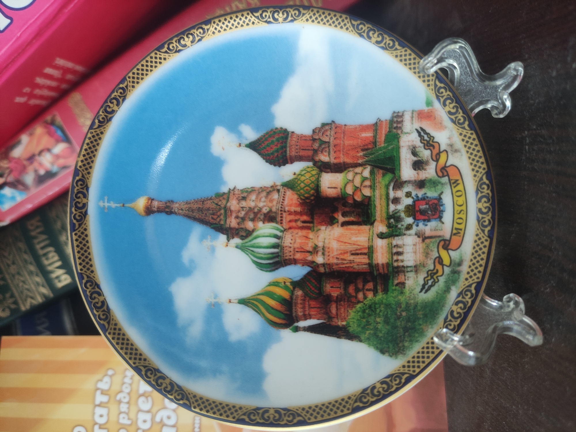 A decorative plate featuring Saint Basil's Cathedral in Moscow is displayed on a dark surface, supported by a clear plastic stand. The plate's rim is adorned with a blue and gold patterned border. In the background, out of focus, are books. One book has the word "БИБЛИЯ" (Bible) visible on its spine. Another book is bright pink. The location is not in Baku, Azerbaijan; the imagery on the plate is of a famous landmark in Moscow, Russia.
