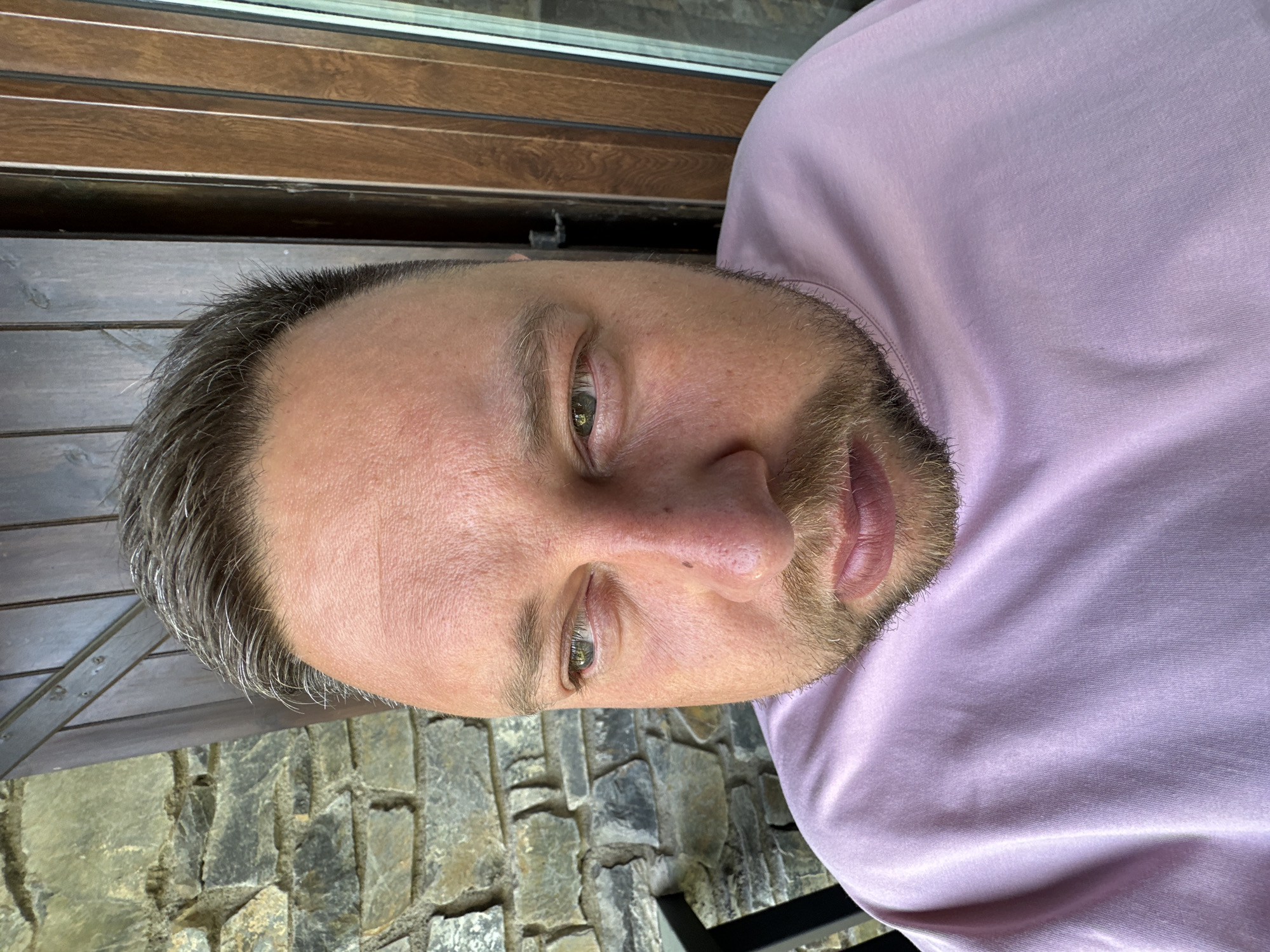 A man is pictured in a close-up portrait. He has a beard and short hair that is graying. He is wearing a light purple shirt. The background appears to be an outdoor setting, featuring a stone wall and wooden accents. The location is possibly Encamp, Andorra.