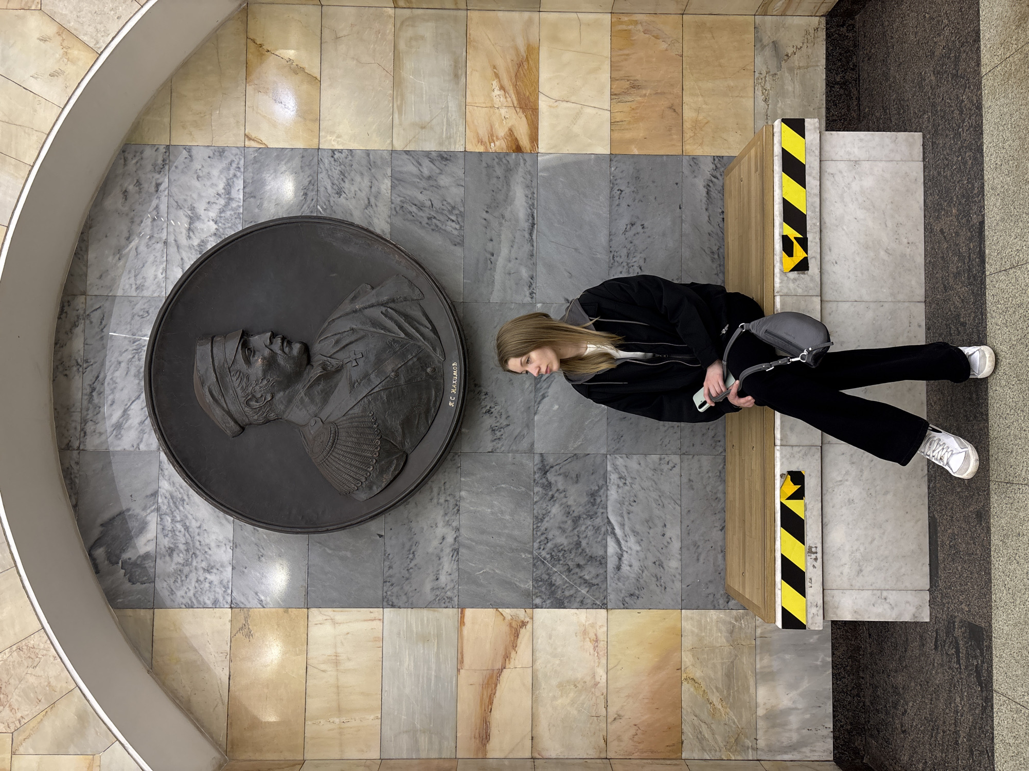 A high-angle shot shows a young woman sitting on a wooden bench next to a large, circular bas-relief in what appears to be an interior space, possibly a subway station or museum, in Saint Petersburg, Russia. The woman has long blonde hair, wears a black jacket, white shirt, black trousers, and white sneakers. She holds a mobile phone. She rests next to a large, dark metal relief of a man's profile, which reads "JF. C. ALEXANDER". The floor is tiled with grey marble near the relief and transitions to tan marble in the upper area. The bench has black and yellow hazard tape. The area beyond the bench features grey tiles. A curved white architectural element frames the top left of the shot.