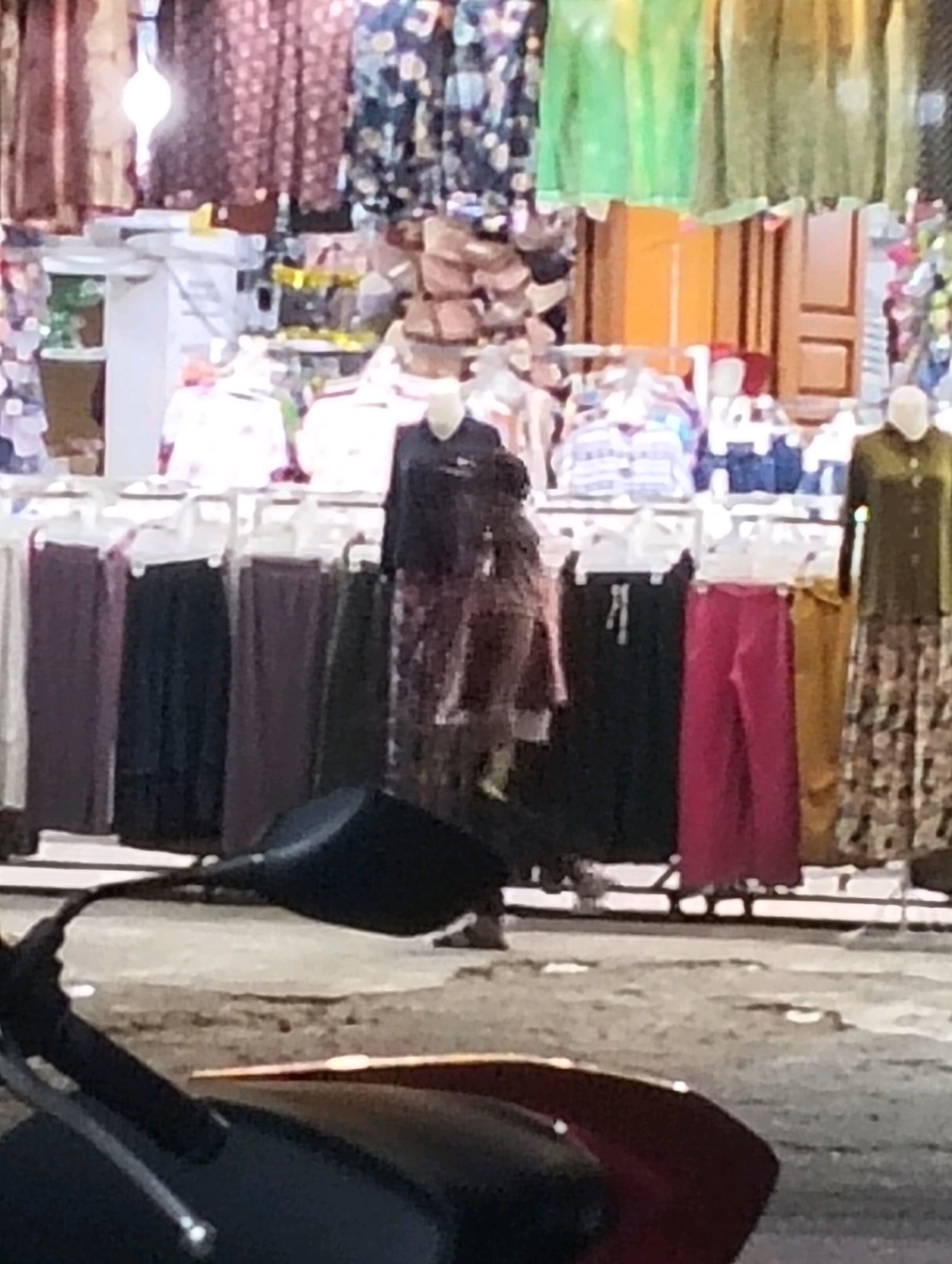 The image depicts an outdoor market stall at night in Grong-Grong, Indonesia. The stall is filled with various garments, including dresses, skirts, and possibly pants, displayed on hangers. Some of the clothing is brightly colored, while other items are darker. In the foreground, parts of a motorcycle are visible, including a handlebar and a side mirror. The ground appears to be paved with a rough texture. The lighting suggests artificial illumination from the stall and possibly surrounding streetlights.