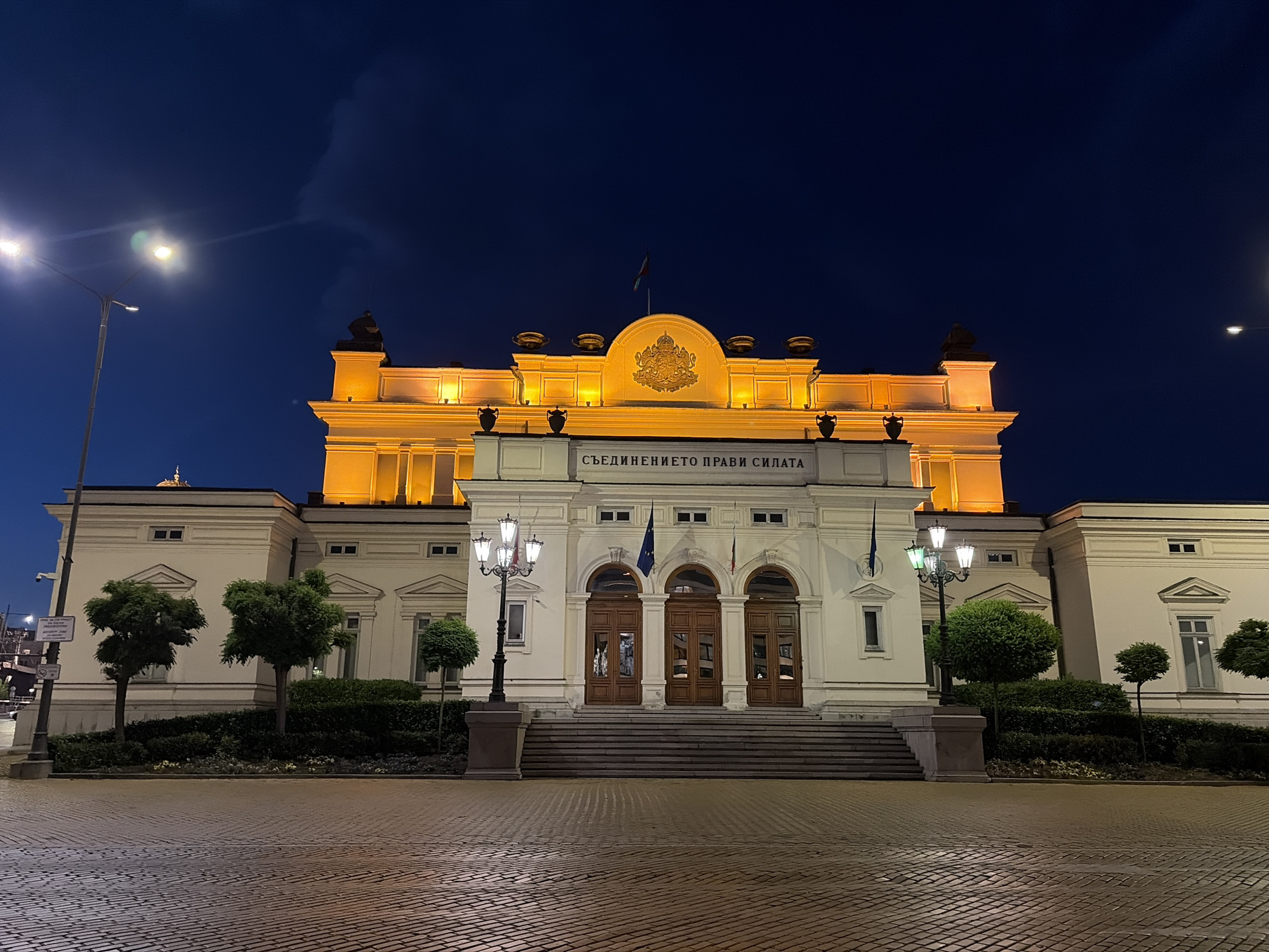 The image depicts the National Assembly of Bulgaria (Parliament) in Sofia, Bulgaria, at night. The sky is a deep indigo blue. The central building, rendered in a classical architectural style, is predominantly off-white or light stone. Its upper facade is brightly illuminated by warm amber lights, highlighting a central semicircular pediment featuring the gold Coat of Arms of Bulgaria and a small Bulgarian flag flying above. Several dark, ornate urn-like structures are visible along the roofline, and dark sculptural elements are present on the upper corners of the illuminated section.
Below the main illuminated section, an inscription in Cyrillic, "СЪЕДИНЕНИЕТО ПРАВИ СИЛАТА" (Union Makes Strength), is prominently displayed. The building's main entrance features three large, arched wooden doors, accessible via a wide, shallow set of stone steps. Two vertical flags, one blue with yellow stars (likely the flag of the European Union) and another tricolor flag (possibly the Bulgarian flag or a similar national flag), hang from poles flanking the central entrance.
The foreground consists of a wide plaza paved with dark, reflective cobblestones, showing highlights from the various light sources. Manicured green hedges and several spherical topiary trees are planted in beds directly in front of the building, separating it from the paved area.
Illumination is provided by several sources: a tall, modern streetlamp with two bright white luminaires stands on the far left, exhibiting lens flare. Two ornamental lampposts, each featuring multiple white lamps, are positioned on stone bases in the mid-ground, flanking the central steps. Another faint streetlight is visible on the far right. A pole with signs and a security camera is visible on the far left. No individuals are present in the scene.