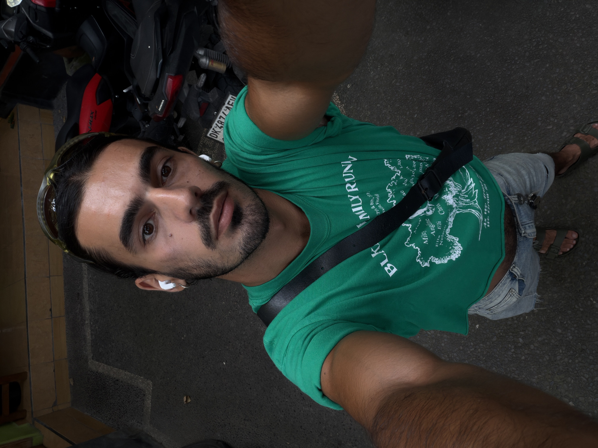 A man is taking a selfie in North Kuta, Indonesia. He is wearing a green t-shirt, denim shorts, and sandals. He has a black strap across his chest and sunglasses on his head. A motorcycle is visible in the background. The license plate number of the motorcycle is DK4874 AEW.
