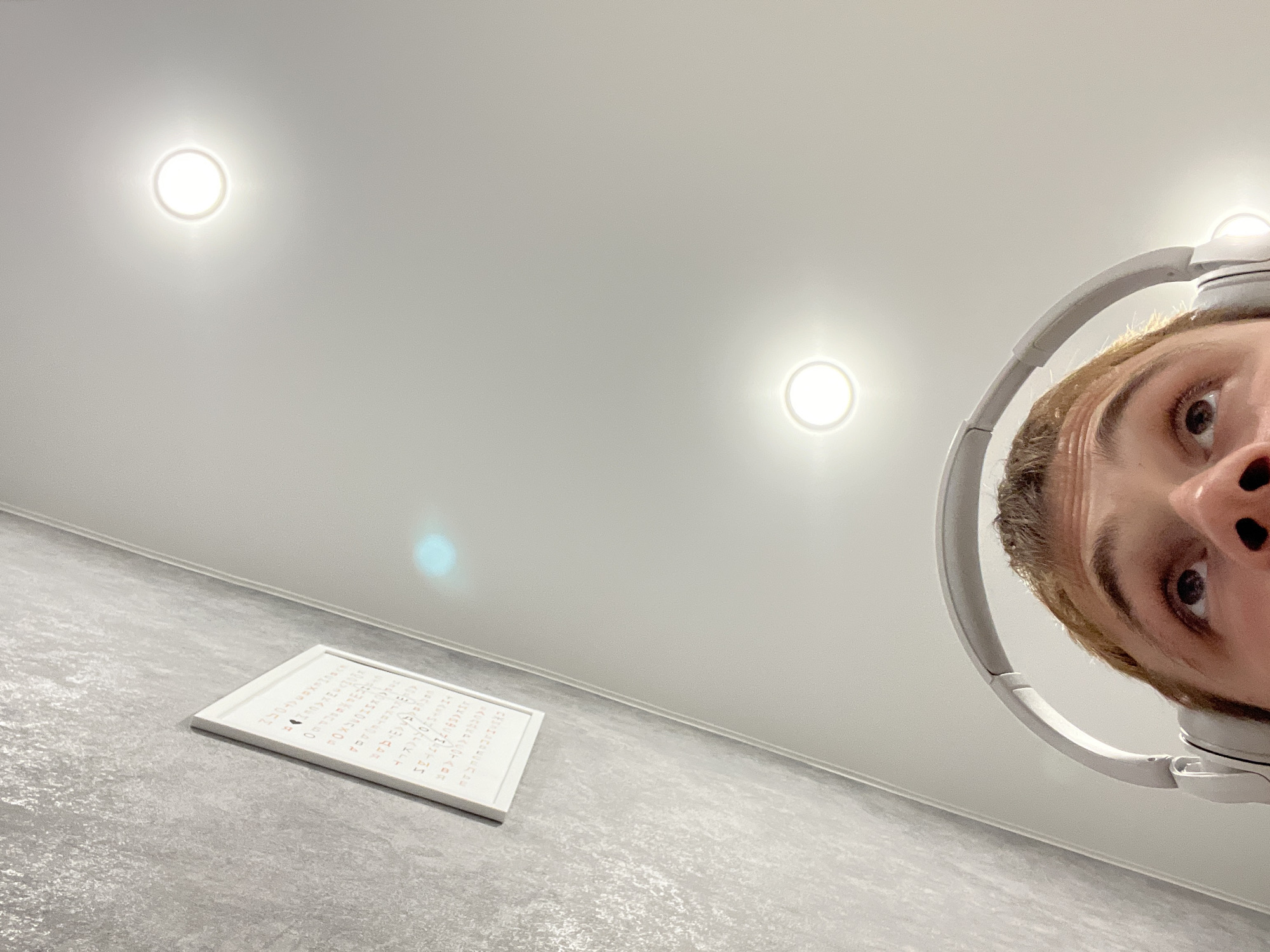 This is a low-angle, wide-shot photograph of a person wearing white headphones. The person's face is visible from the eyes upwards, positioned in the upper right quadrant of the frame. Their eyes are wide and looking towards the camera. The image appears to be taken indoors, possibly in a room with a light-colored, textured floor and a white ceiling. Two circular lights are visible on the ceiling, casting a bright glow. In the lower left quadrant of the image, a white rectangular object with text arranged in columns is placed on the floor. The text appears to be in a foreign script, possibly Japanese or Chinese characters. The overall lighting is bright, suggesting it could be daytime or that the room is well-lit. The composition is somewhat abstract due to the extreme low angle and the cropped view of the subject.